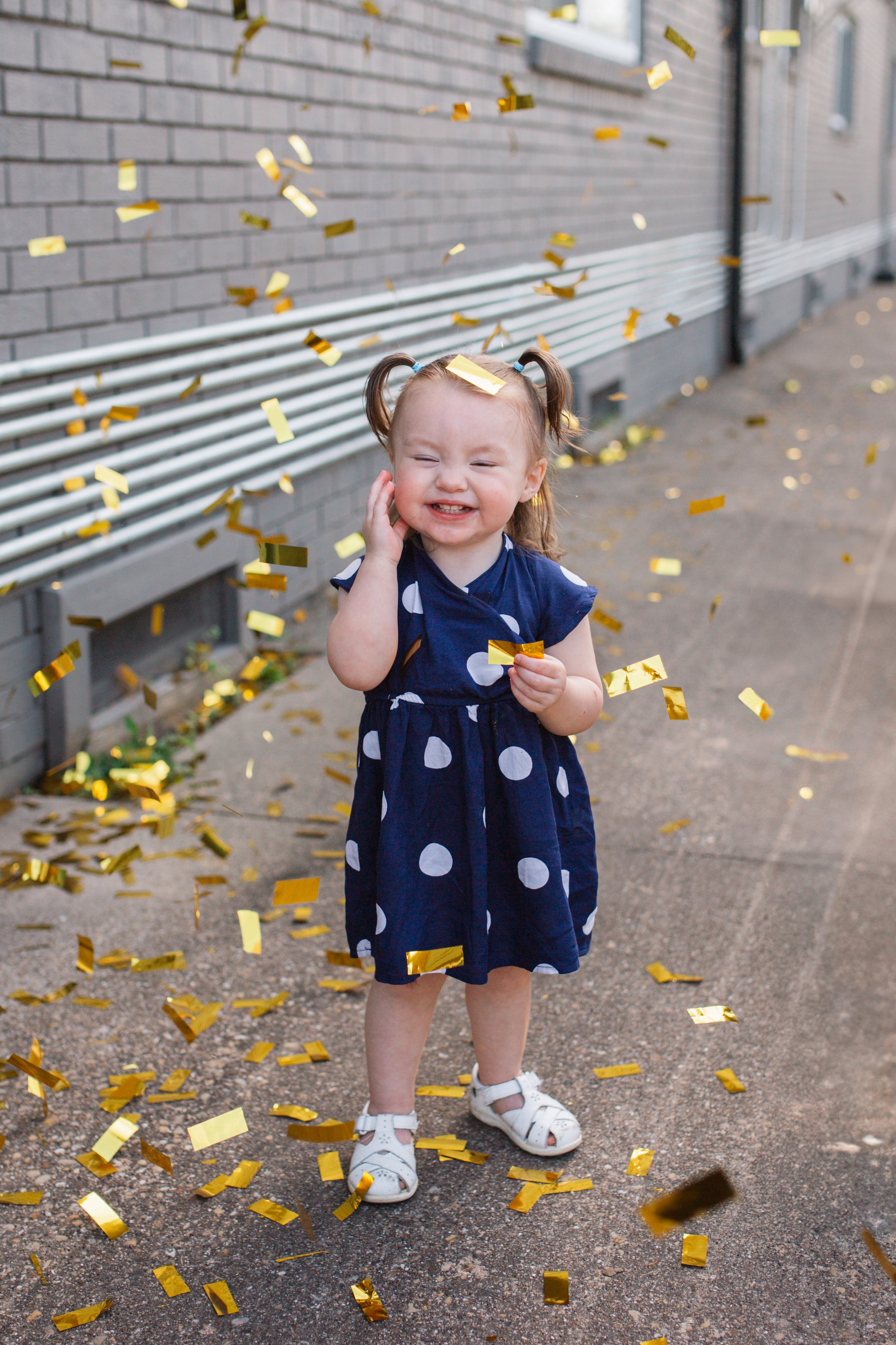 A young girl with pigtails and a big smile under falling gold confetti, standing outdoors in front of a brick building and metal pipes.