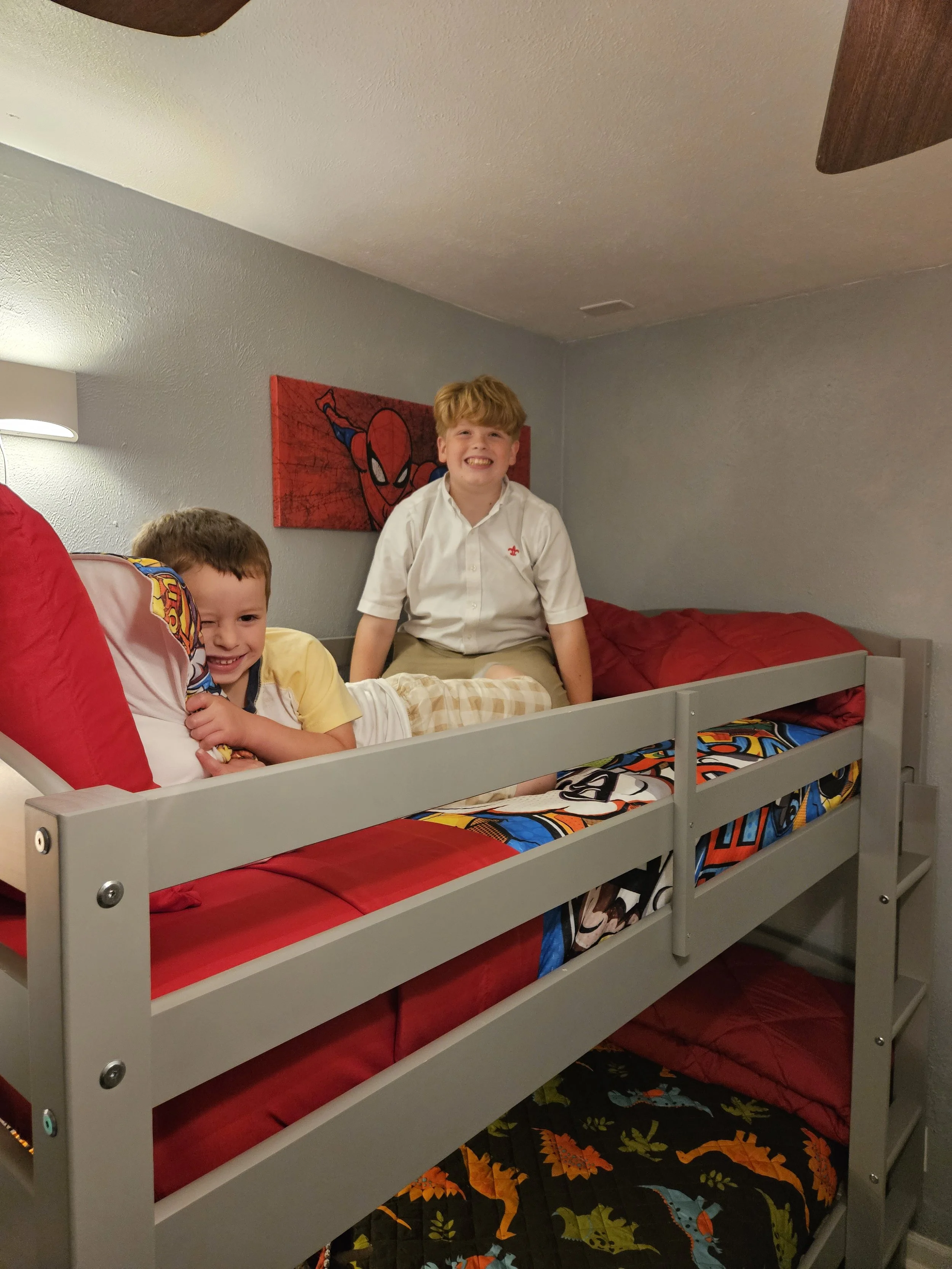 Two boys sitting on a bed and smiling in a child's bedroom. The bed has red and colorful superhero-themed bedding. There is a Spider-Man picture on the gray wall behind them.