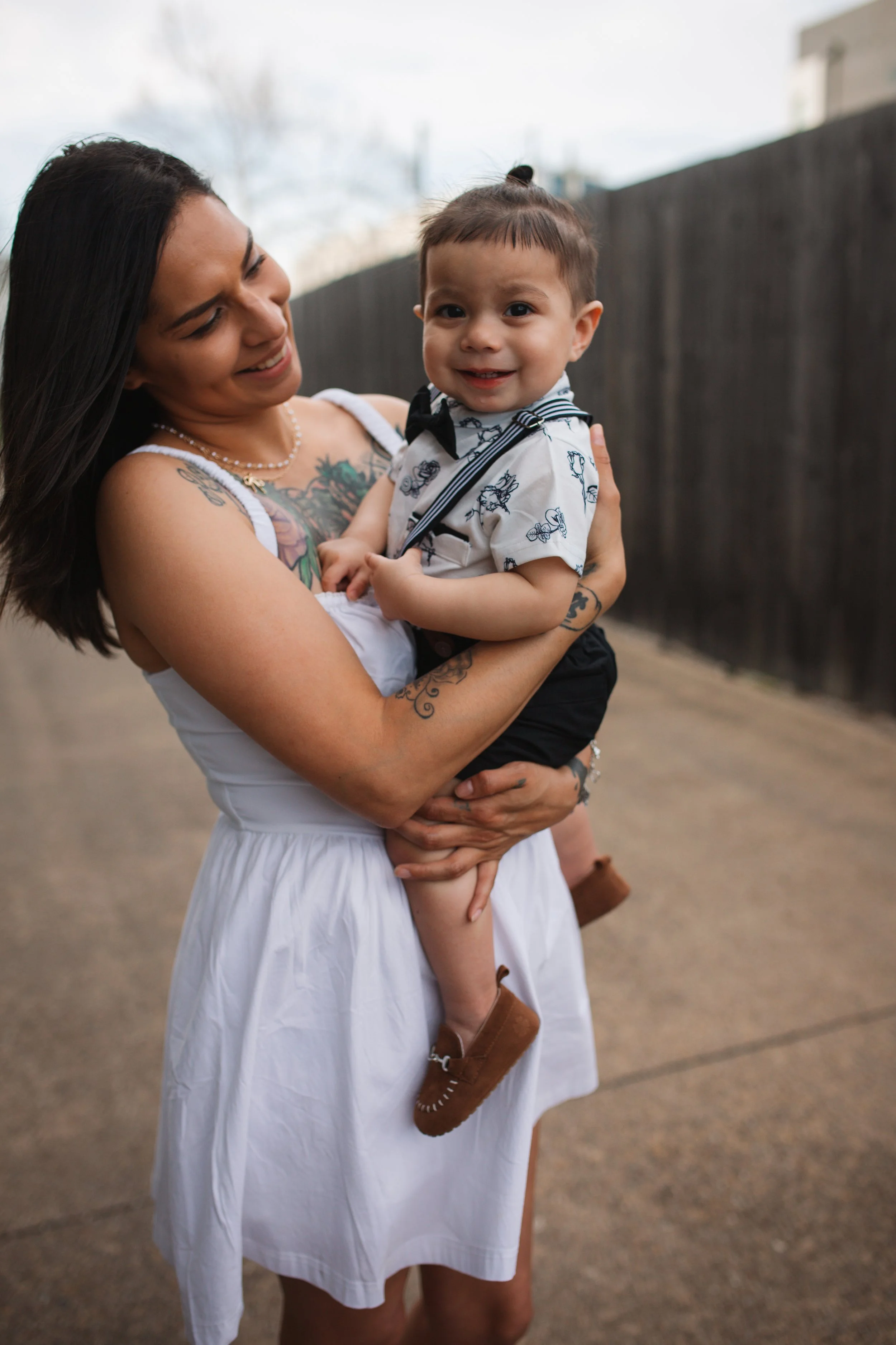 A woman with long dark hair and tattoos on her arm is holding a young boy dressed in a white shirt with a black bow tie. They are standing outdoors on a dirt road with a black fence and some buildings and trees in the background. The woman is smiling at the boy, who is smiling directly at the camera.