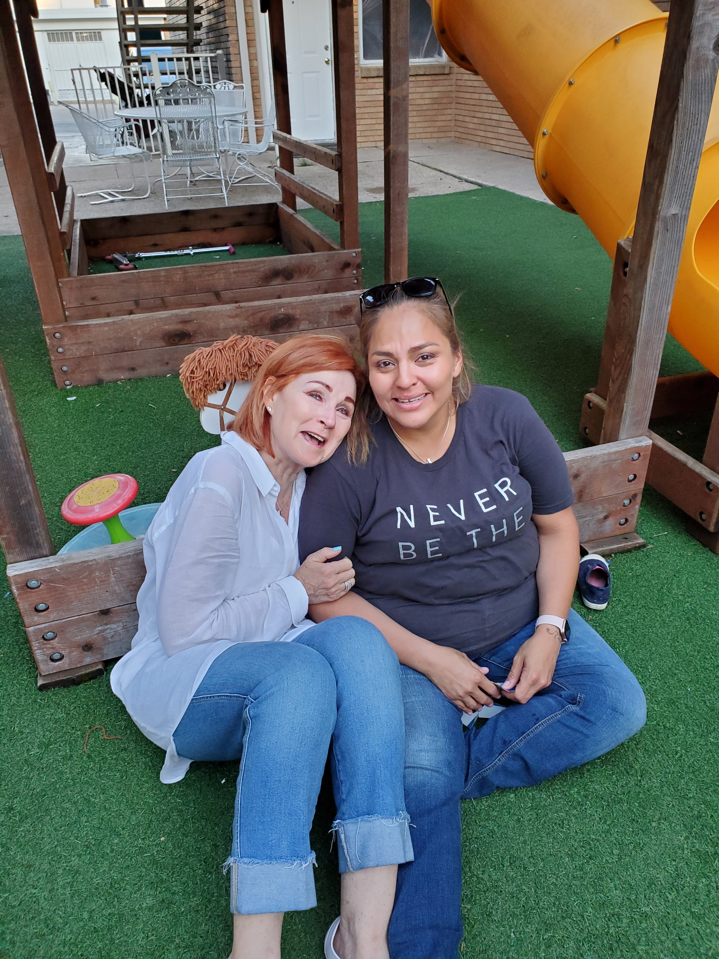 Two women sitting close together under a wooden play structure with a yellow slide in a backyard. One woman has short red hair and is wearing a white shirt, while the other woman has long brown hair, sunglasses on her head, and a dark t-shirt. They are smiling and appear happy.