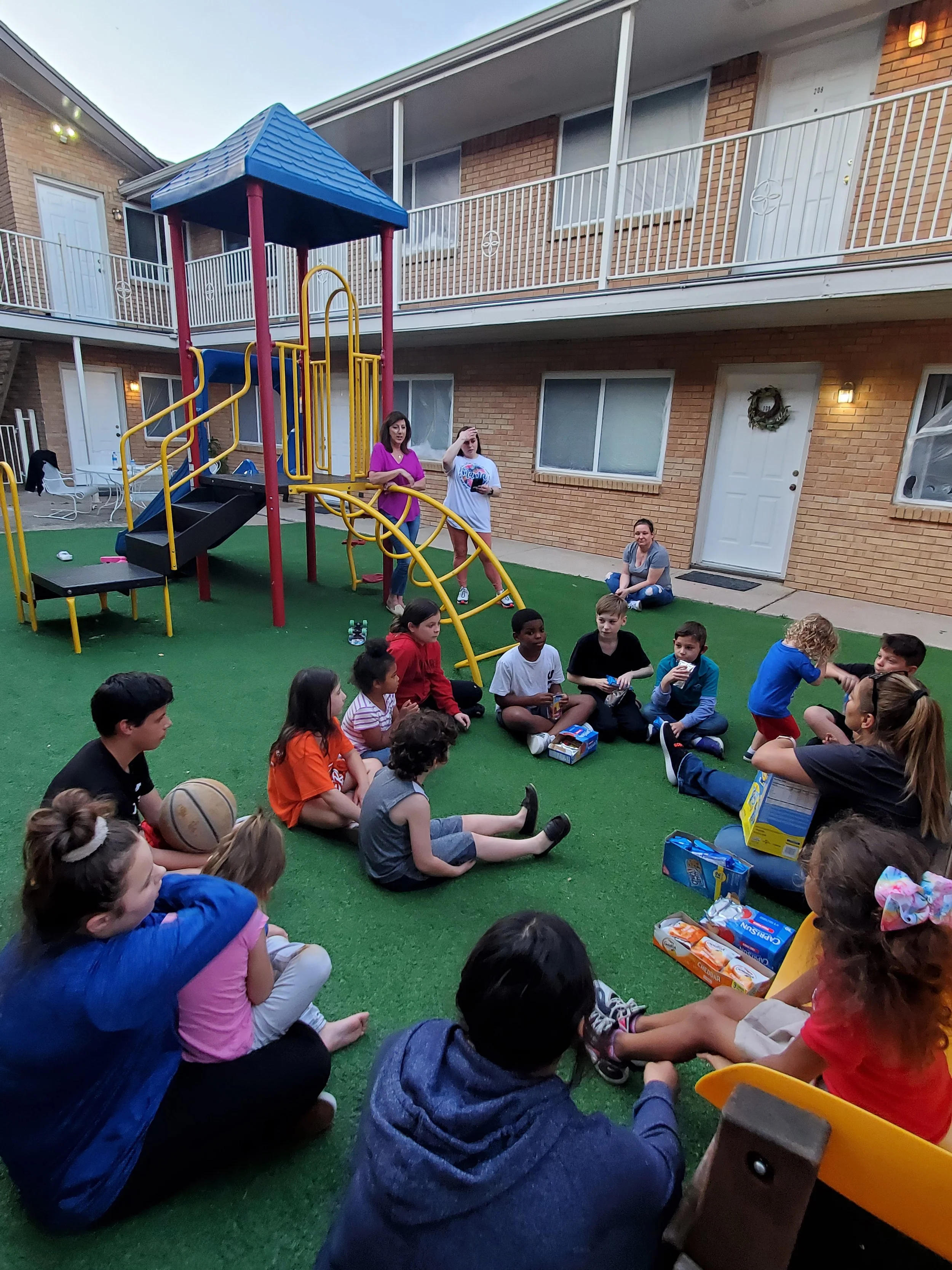 Group of children and adults sitting on green artificial turf in an outdoor area, with a playground structure and a brick apartment building in the background. Some children hold snacks or drinks, and a woman stands near the playground equipment.
