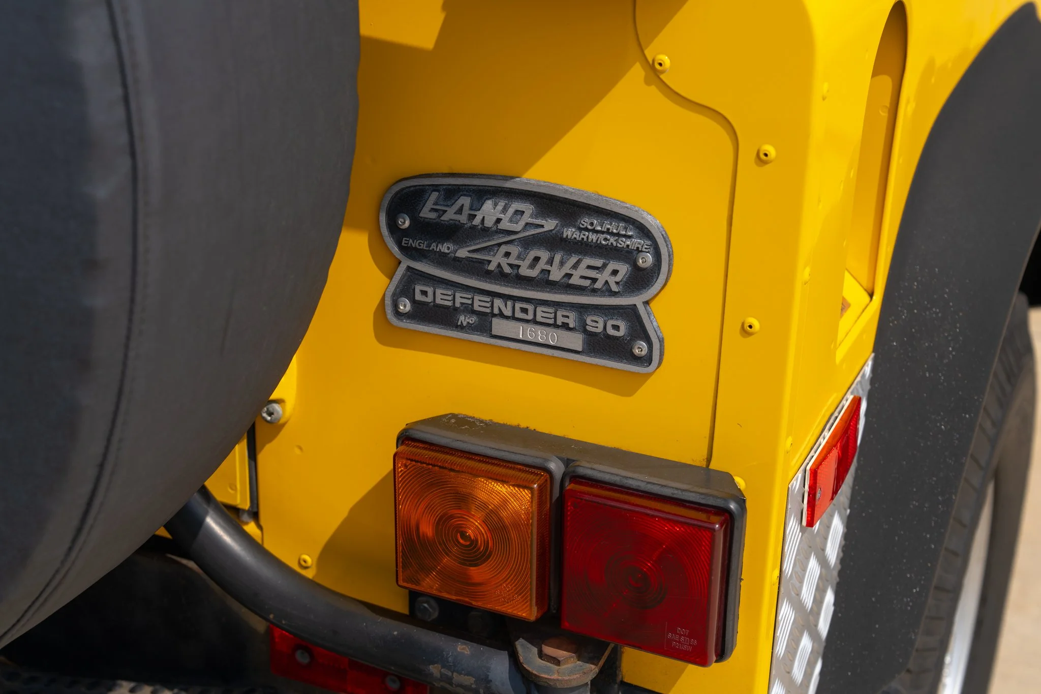 Close-up of the rear side of a yellow Land Rover Defender 90, showing a metallic badge with the Land Rover logo and model name, along with tail lights.