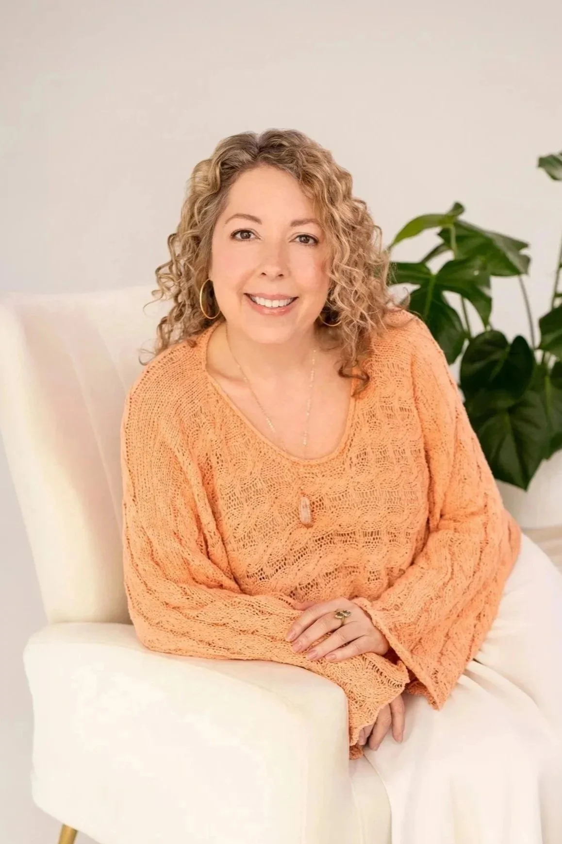 Jenic, the Founder of Little Lightworkers. A woman with curly blonde hair, wearing an orange lace top, sitting on a white chair, with a large green plant behind her, smiling at the camera.