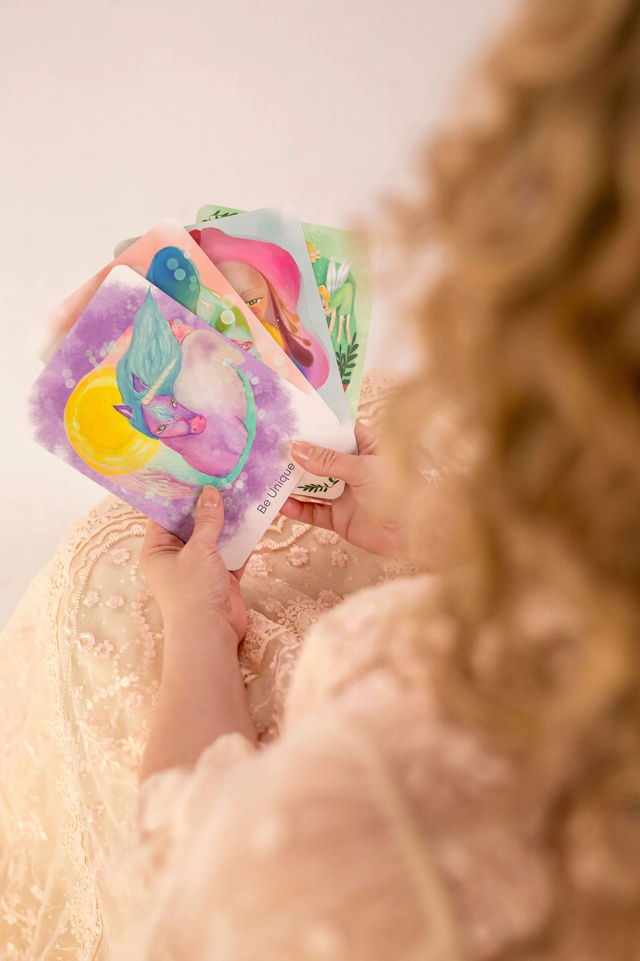 A person with curly blond hair in a lace dress is holding colorful illustrated postcards with fantasy characters, including a unicorn, in a bright setting.