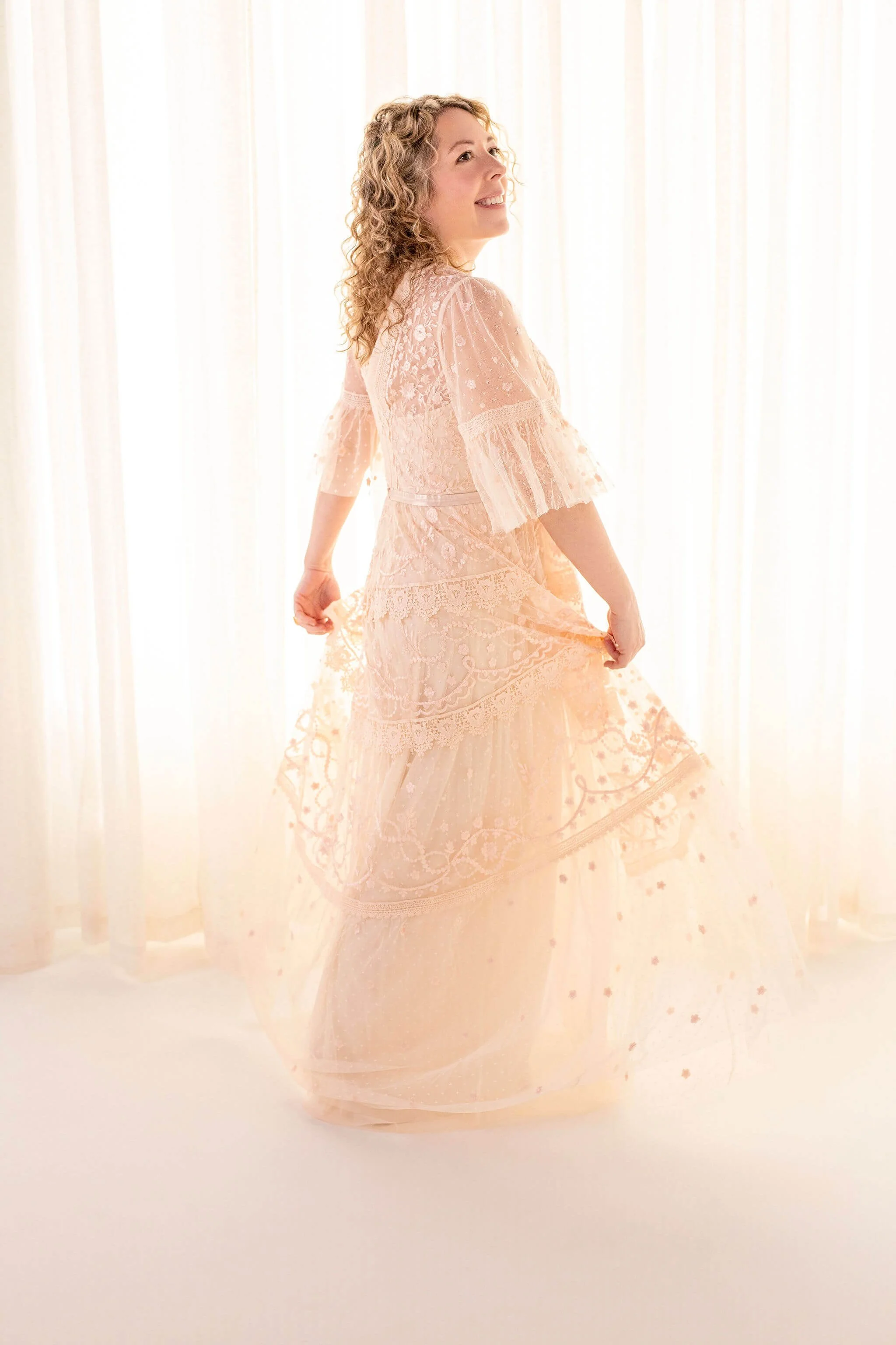 Jenic, the Founder of Little Lightworkers. Woman in a peach lace dress with puffed sleeves, standing in front of light-colored curtains, smiling and holding the hem of her dress.