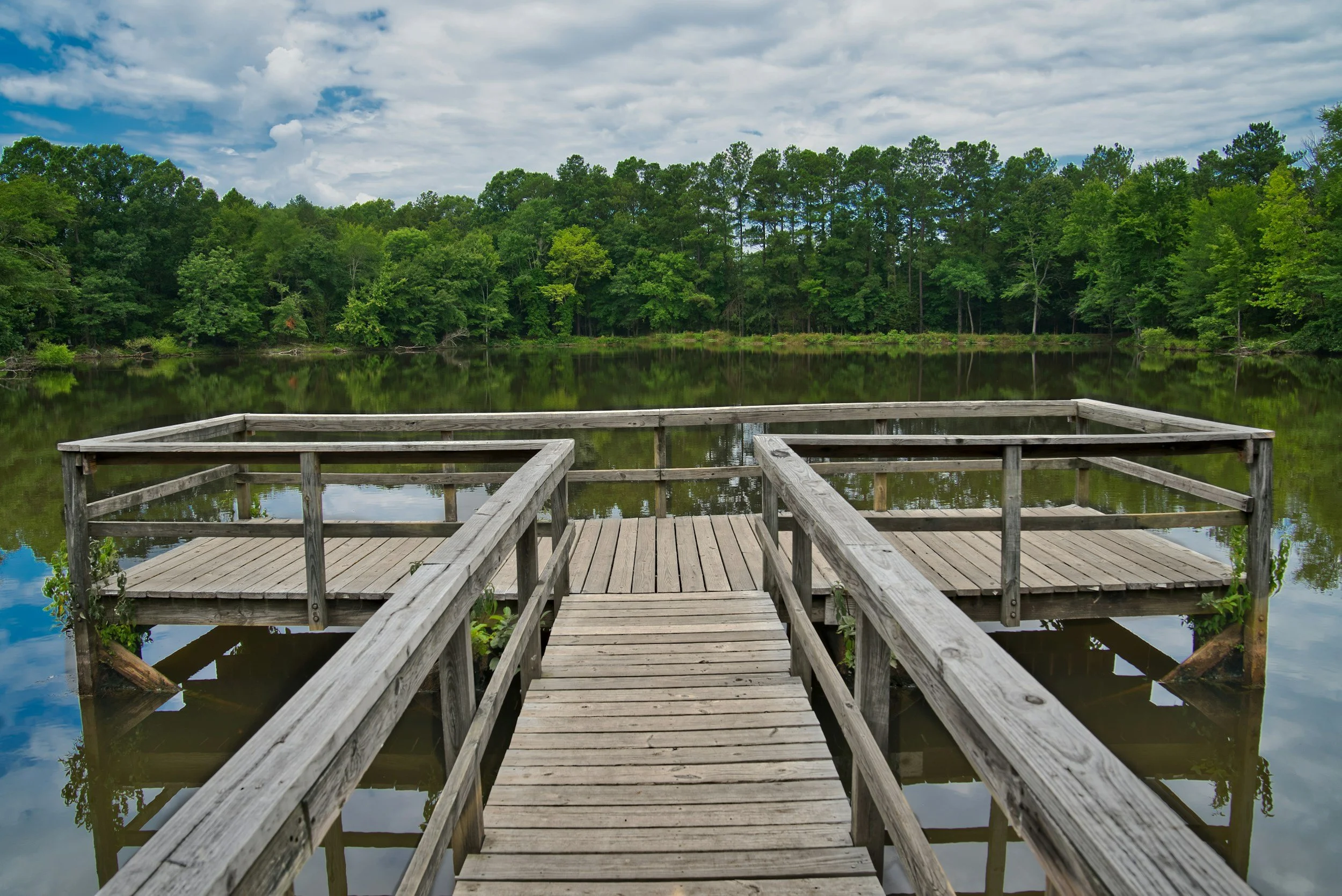 A wooden dock extends into a calm lake surrounded by lush green trees, under a partly cloudy sky.