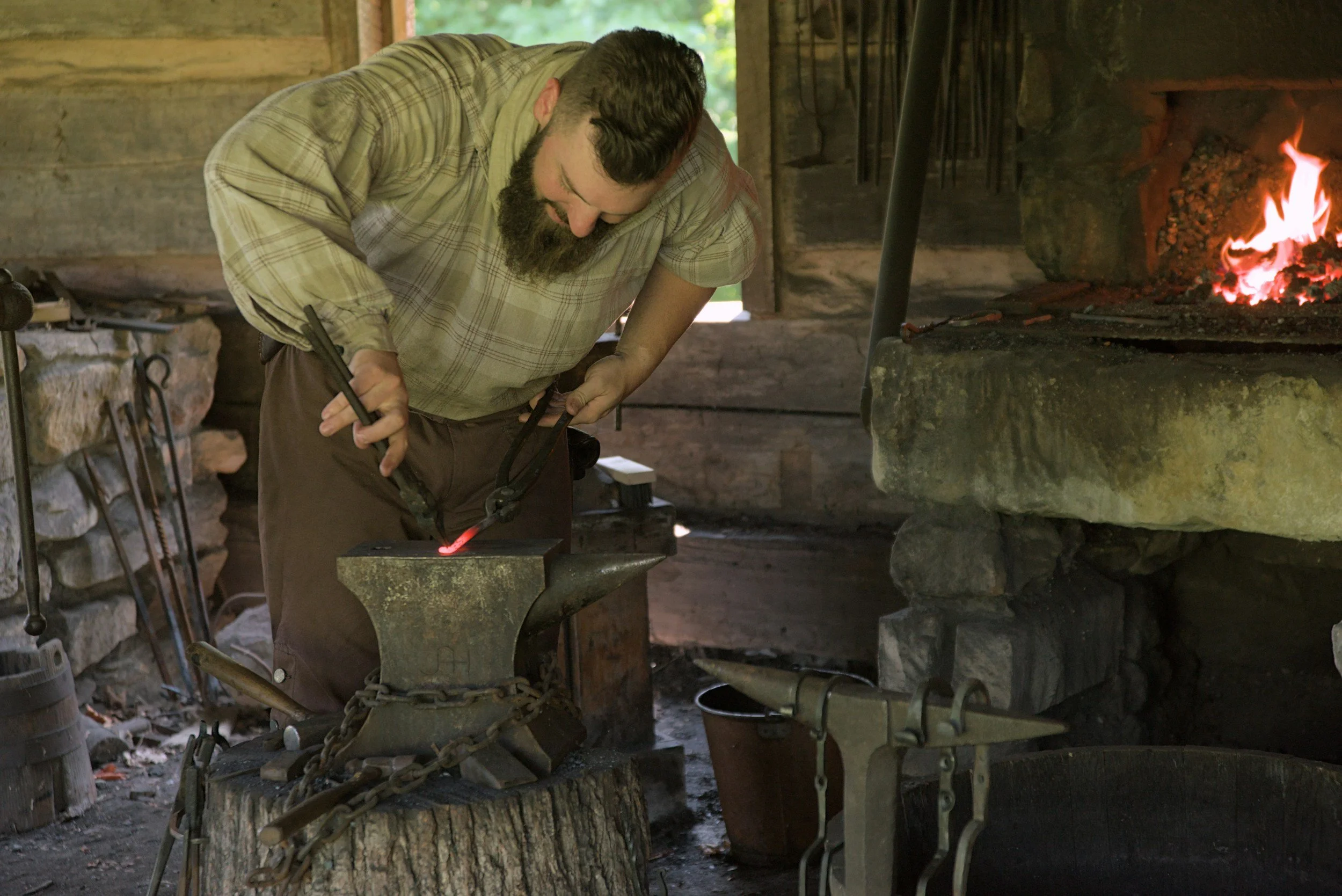 A blacksmith with a beard working at an anvil in a rustic blacksmith's workshop, using tongs to hold a piece of heated metal near a forge.