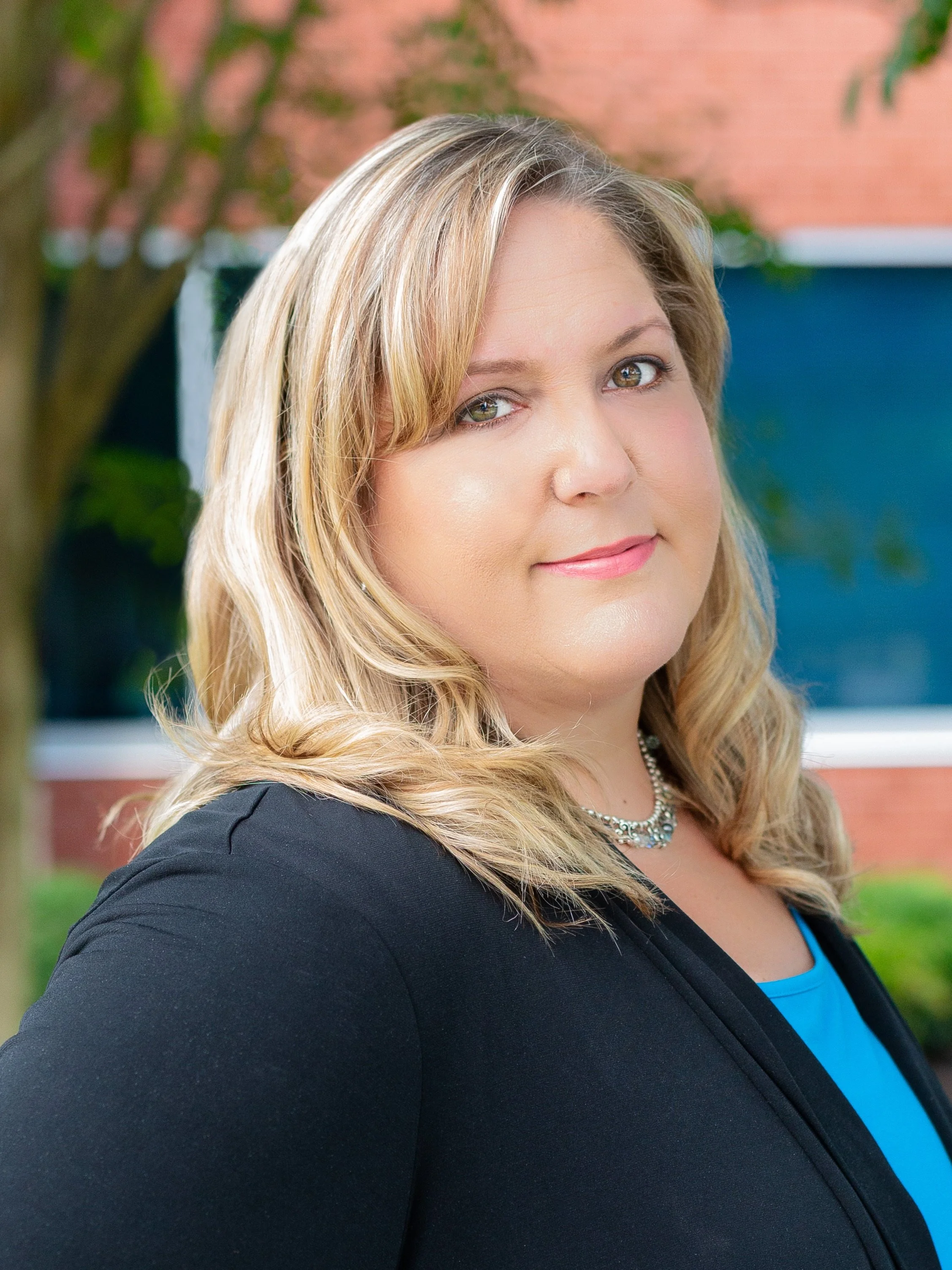 A woman with blonde hair, wearing a black blazer and a blue top, standing outdoors near a brick building with trees in the background.