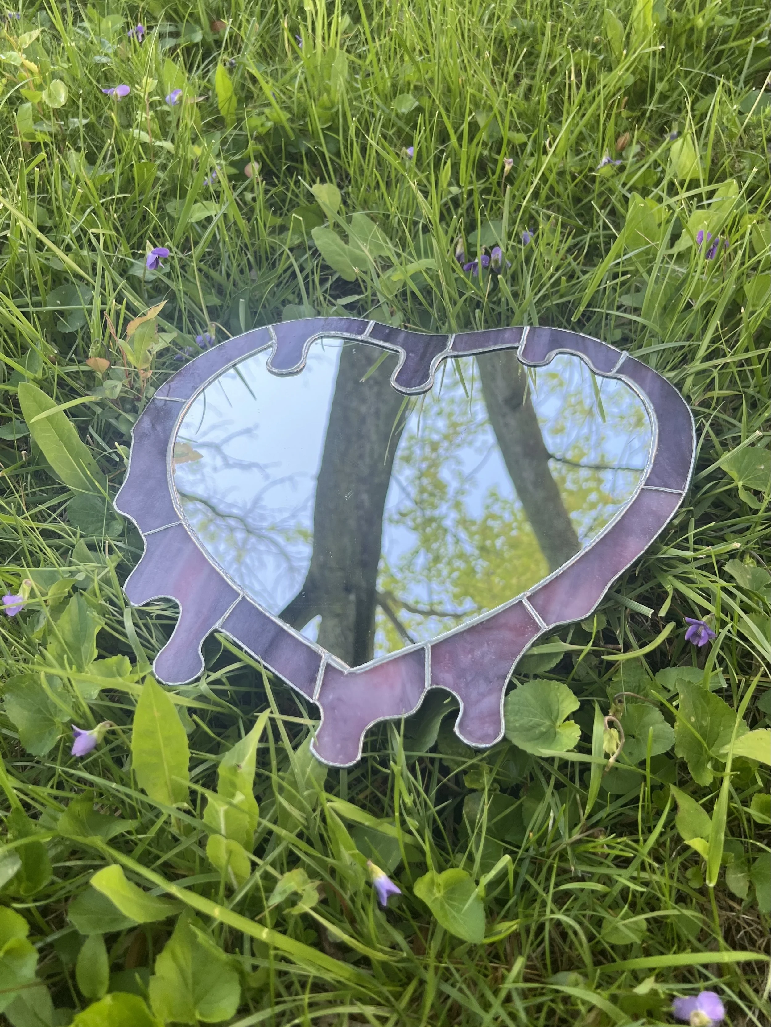 A heart-shaped mirror with a reflective surface, surrounded by a puzzle-piece border, laying on green grass with small purple flowers, reflecting blue sky and tree branches.
