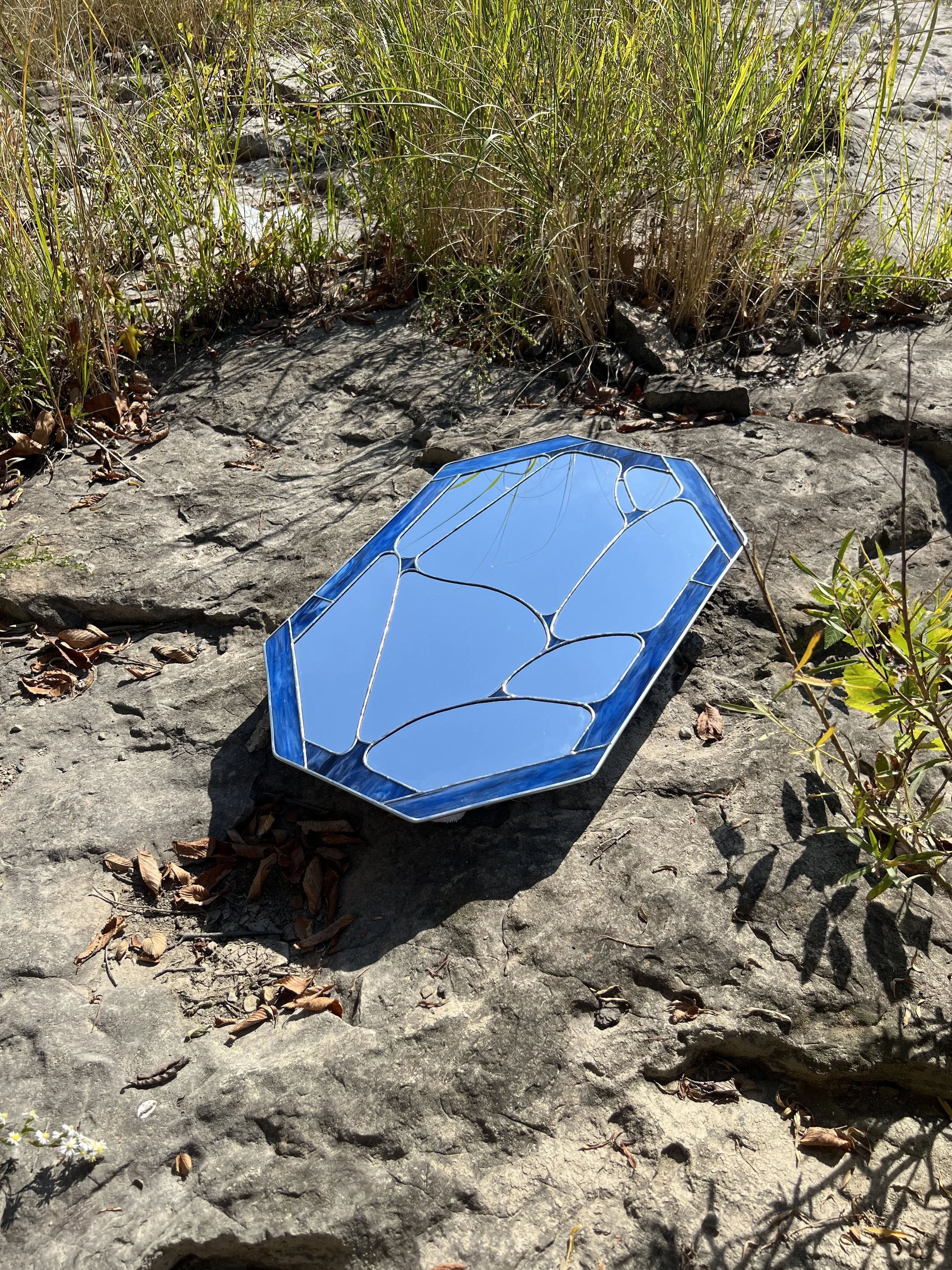 A decorative mirror in a geometric, multi-sided shape with a blue border, lying on a rocky surface outdoors near grass and plants.