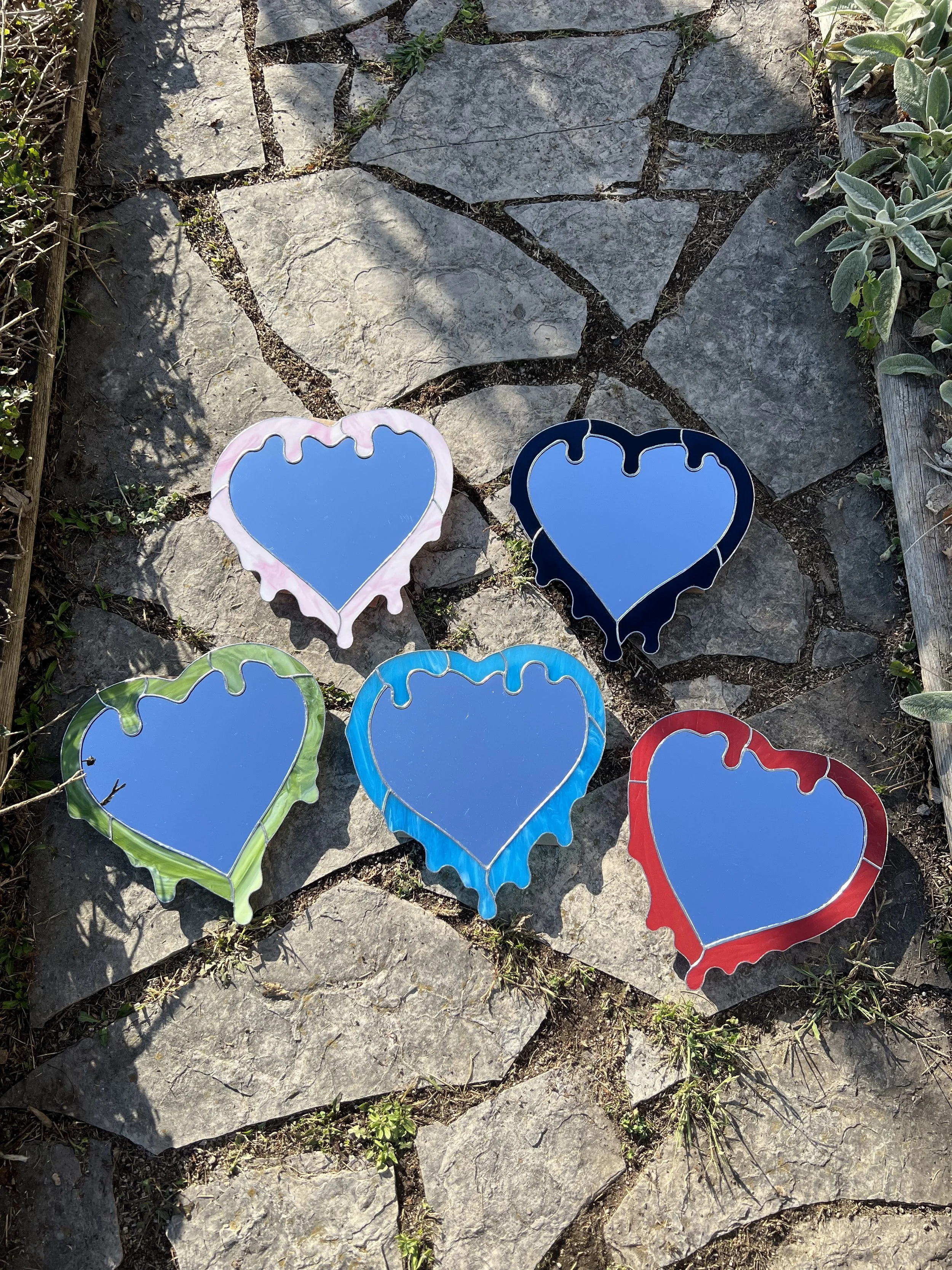 Five colorful heart-shaped mirrors with dripping designs on a stone pathway, surrounded by small plants.
