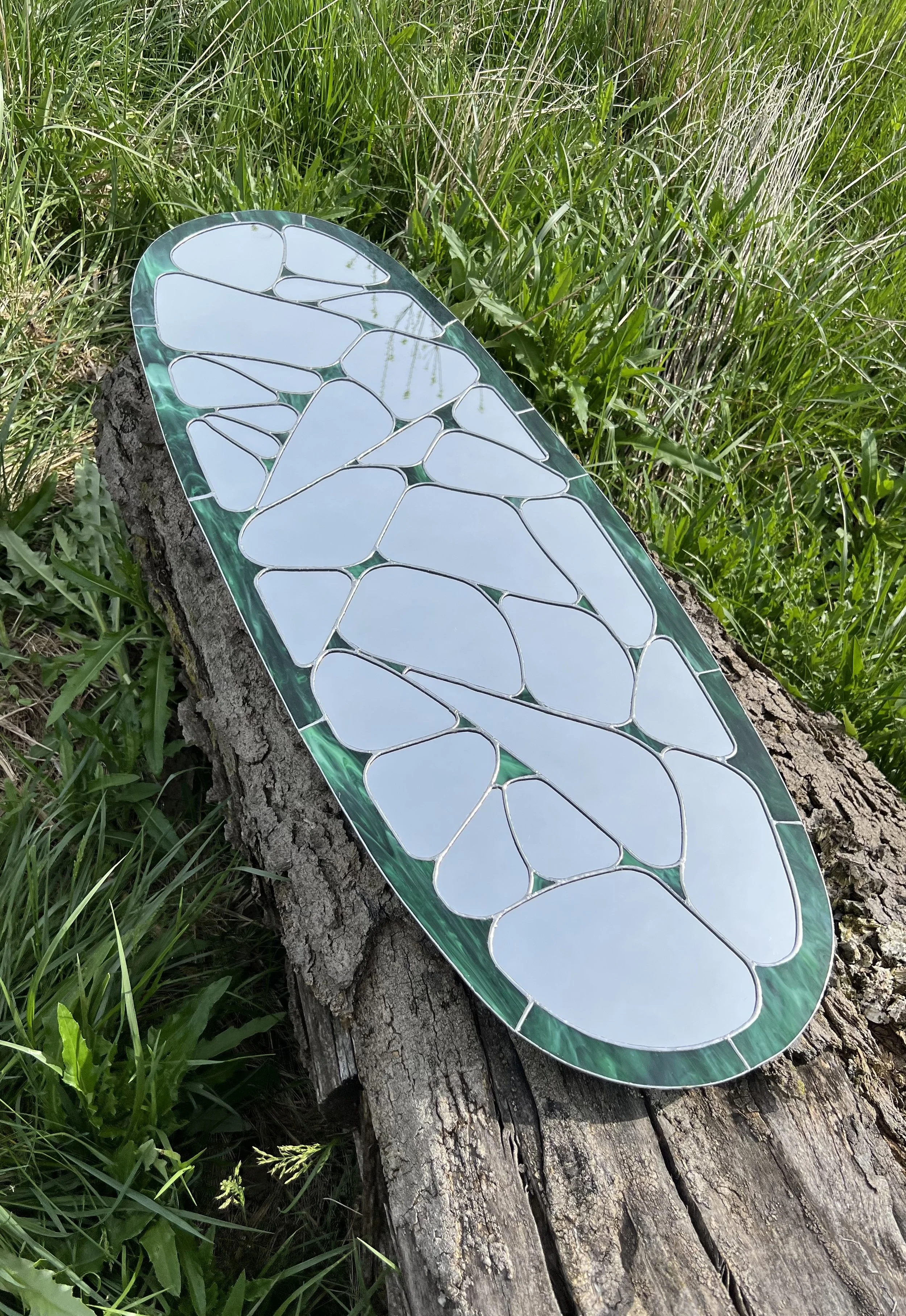 A decorative mirror with irregular, mosaic-like glass pieces in white and green, placed on a fallen tree trunk in a grassy outdoor area.