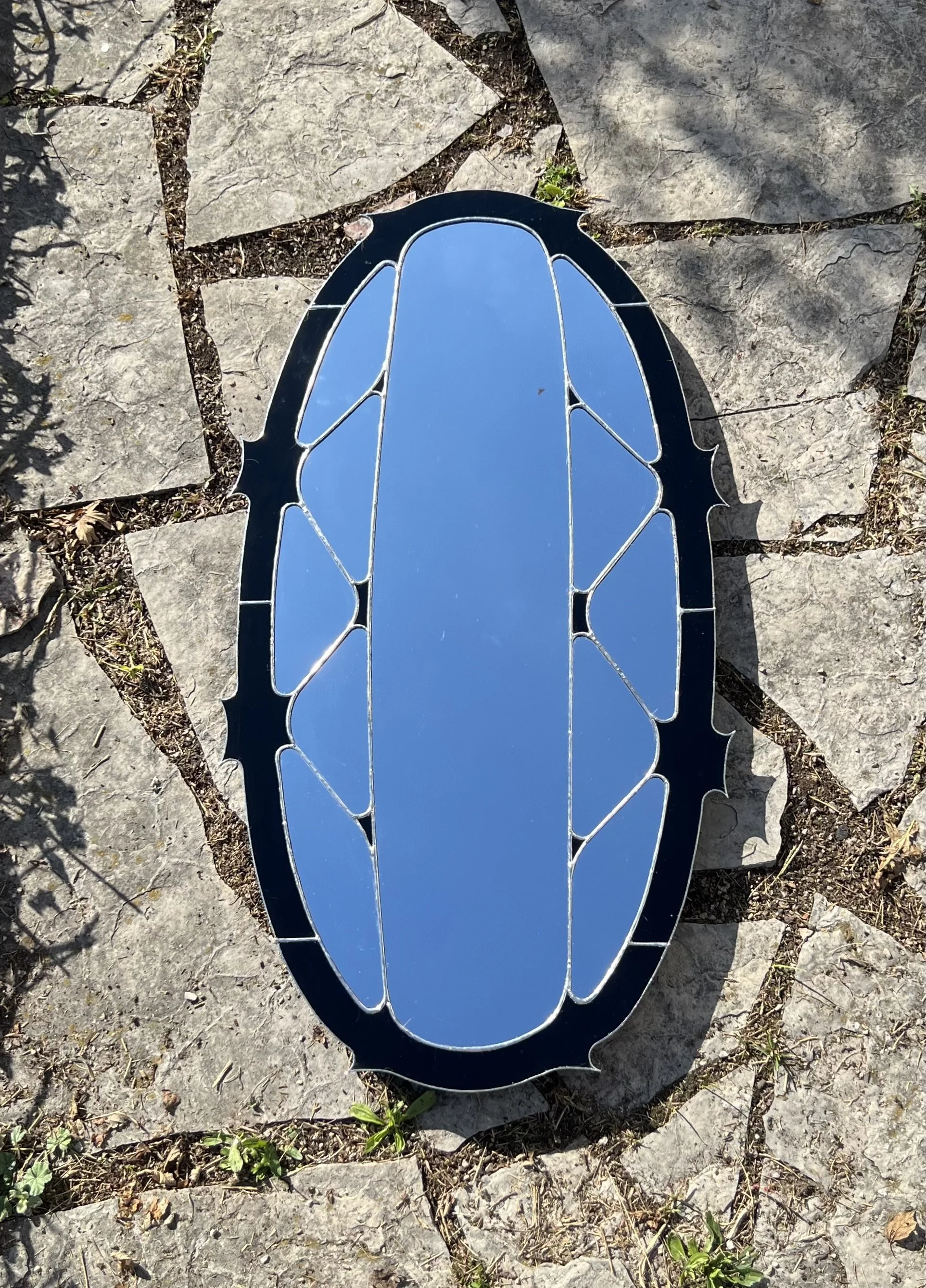 A decorative mirror with a black frame featuring a jagged, crown-like design, placed on a stone surface with small plants and dirt visible around it.
