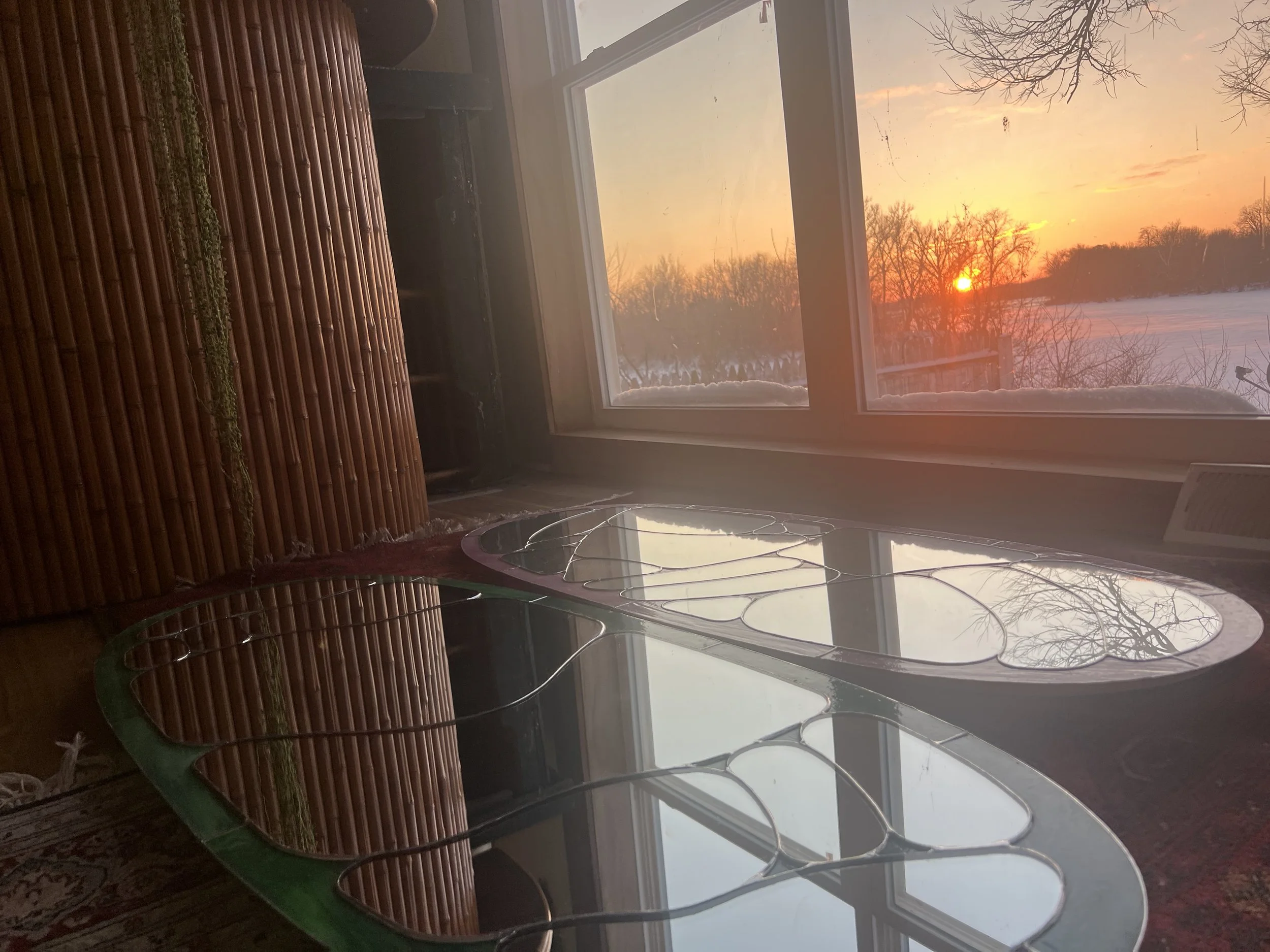 Indoor scene with a large window showing a winter landscape at sunset, with snow-covered ground and leafless trees, and a glass-topped table with wire decorative accents in front of the window.