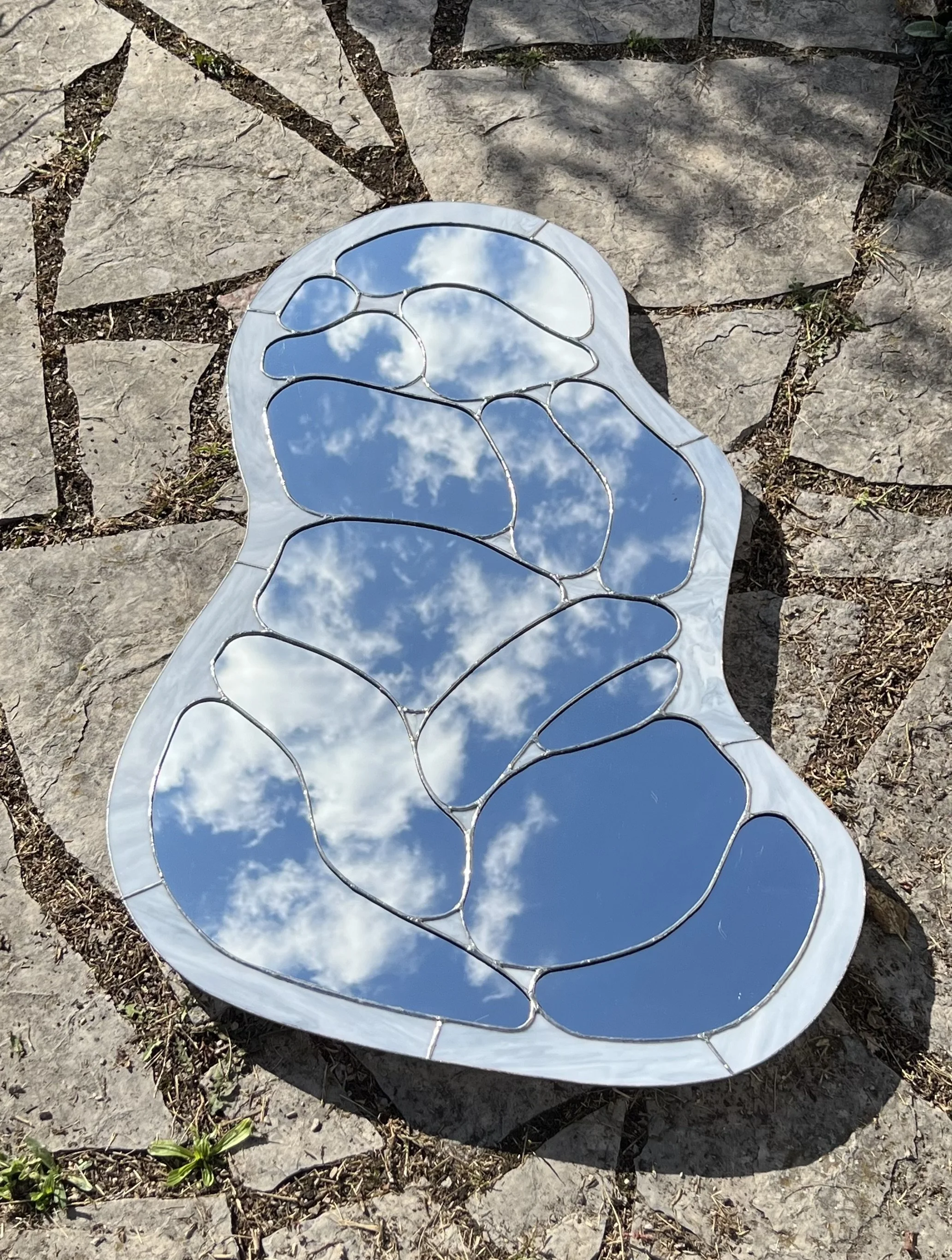 A decorative mirror with a fragmented or stained glass design, reflecting the sky with clouds, placed on a stone surface outdoors.