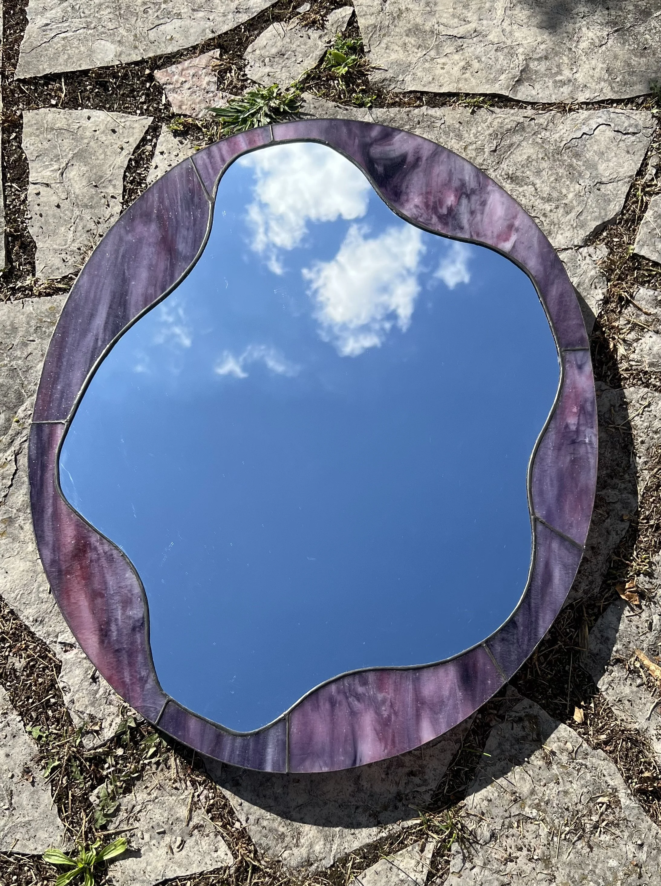Reflection of blue sky and white clouds in an irregularly shaped mirror with a purple and pink marble border, placed on rocks and grass.