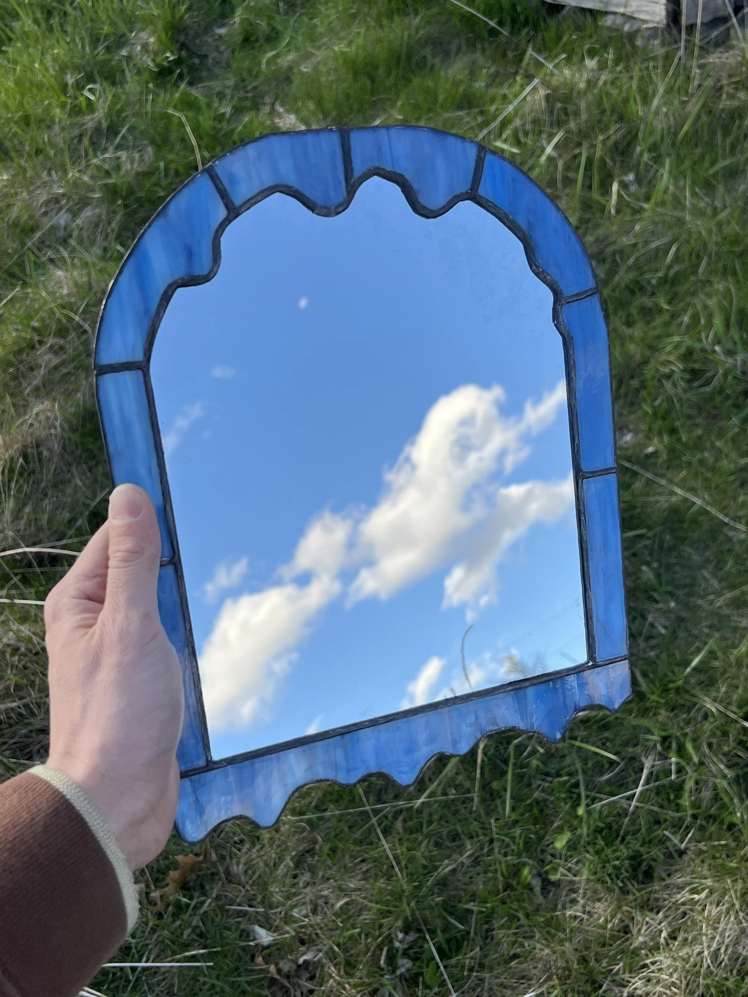 A hand holding a mirror with a blue frame, reflecting a blue sky with clouds.