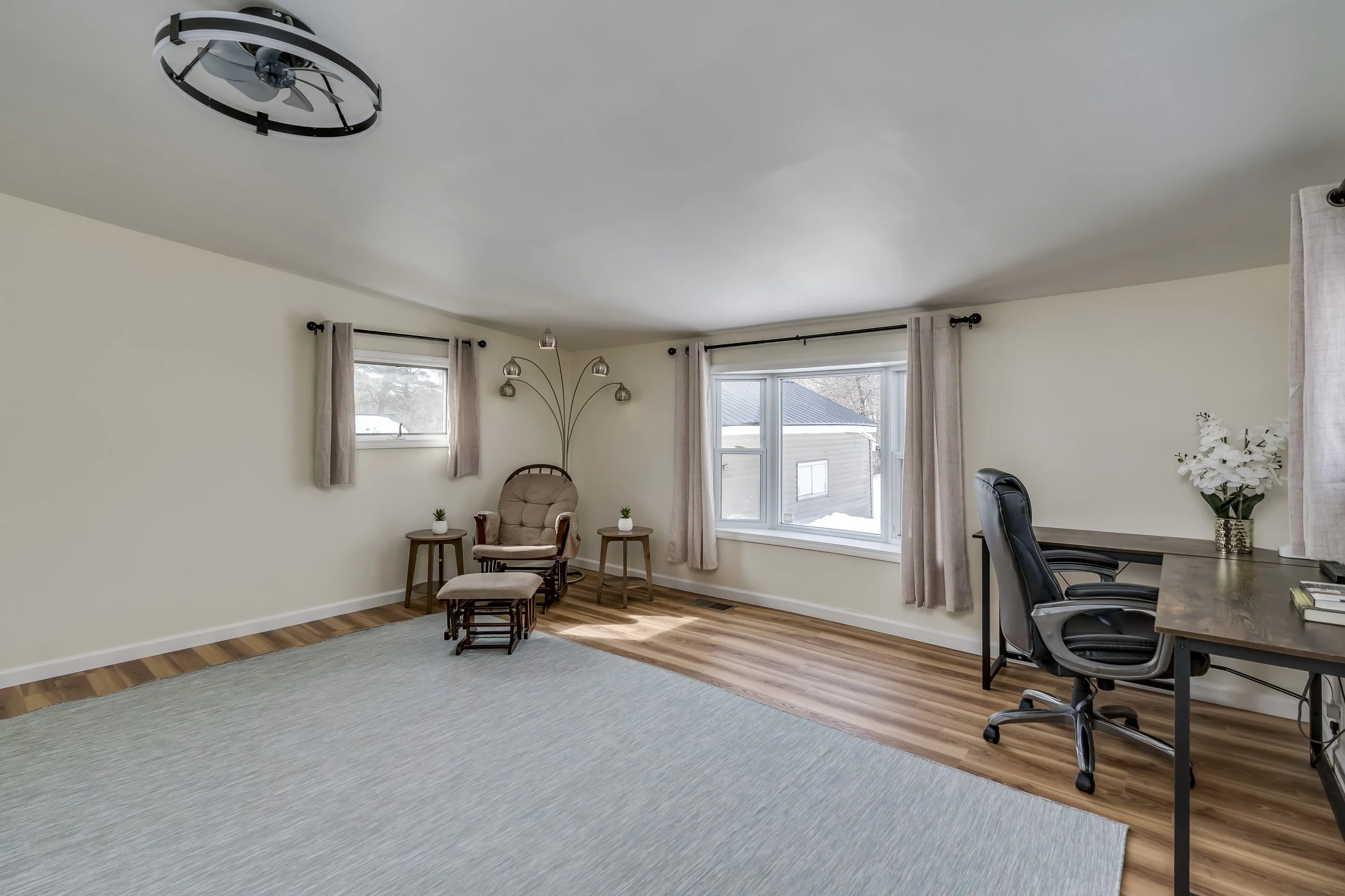 Living room with beige walls, hardwood floors, a large window with light curtains, a small window, beige armchair with footrest, small side tables with plants, a black office chair at a wooden desk, and a ceiling light fixture.