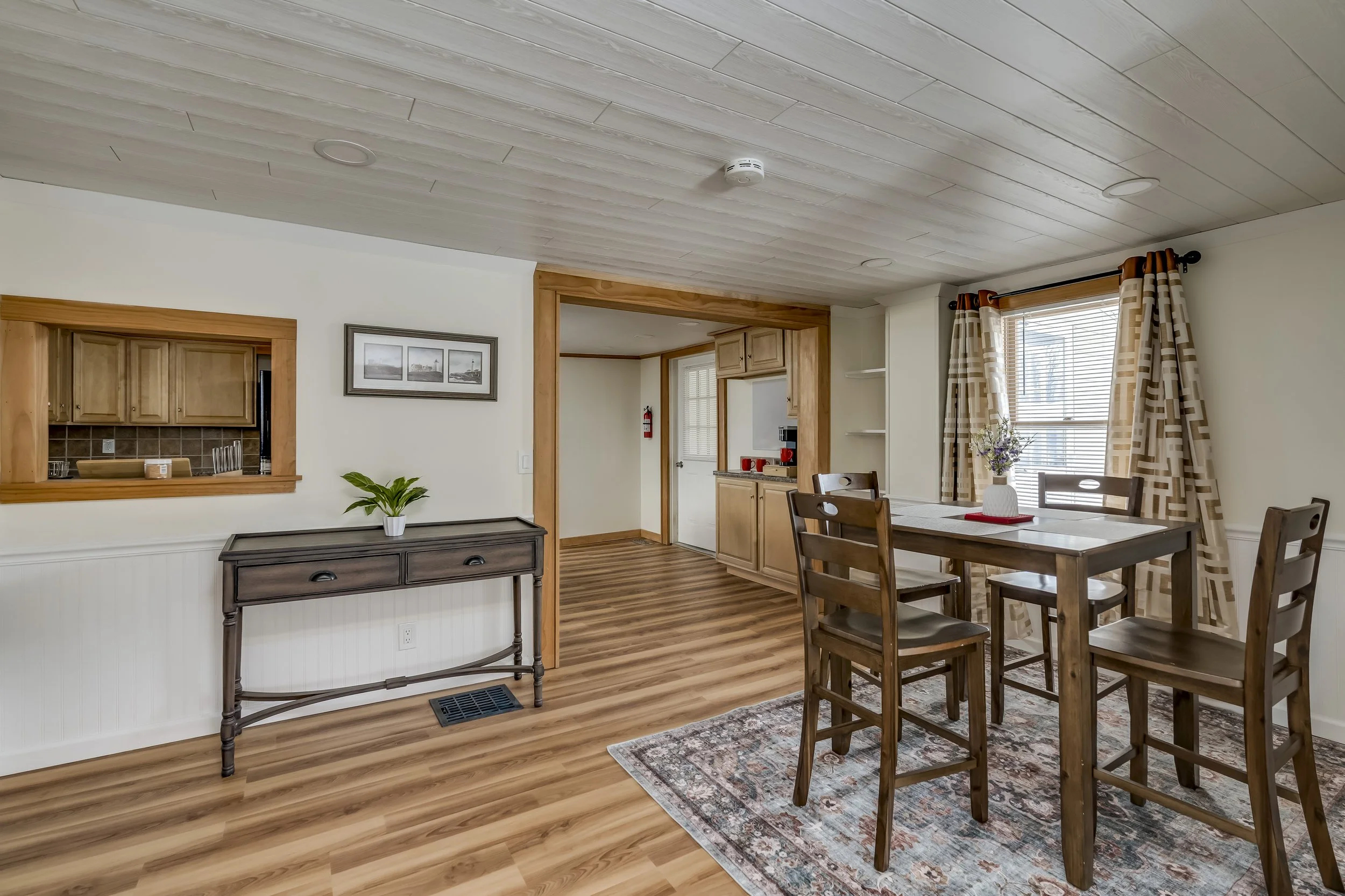 Dining room with wooden table and chairs, window with patterned curtains, and view into kitchen with wooden cabinets.
