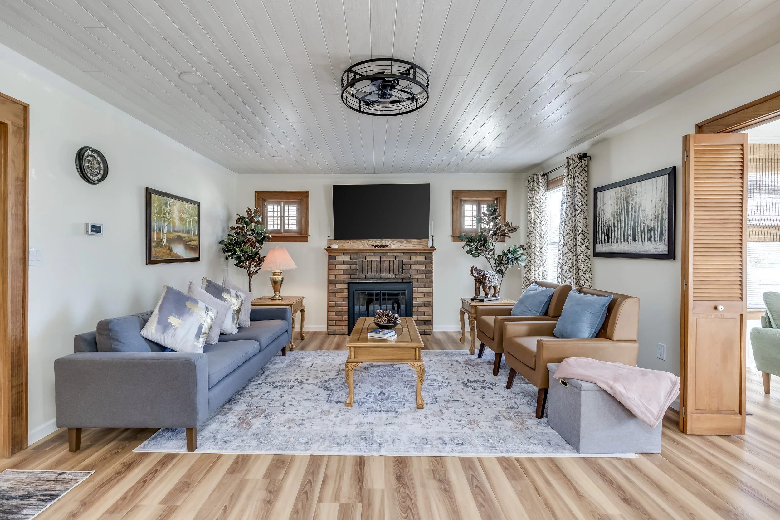 Living room with a brick fireplace, wall-mounted TV, wooden coffee table, gray sofa, two brown armchairs, side tables with lamps and plants, framed paintings, and hardwood floors.