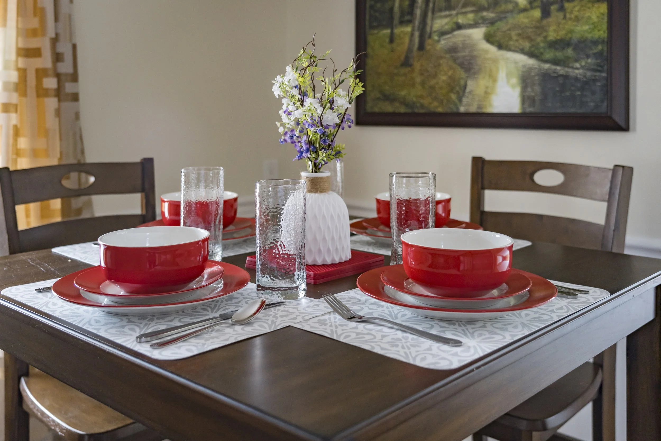Dining table set with red and white dishes, drinking glasses, silverware, and a white vase with purple, white, and green flowers, in a room with a landscape painting on the wall.