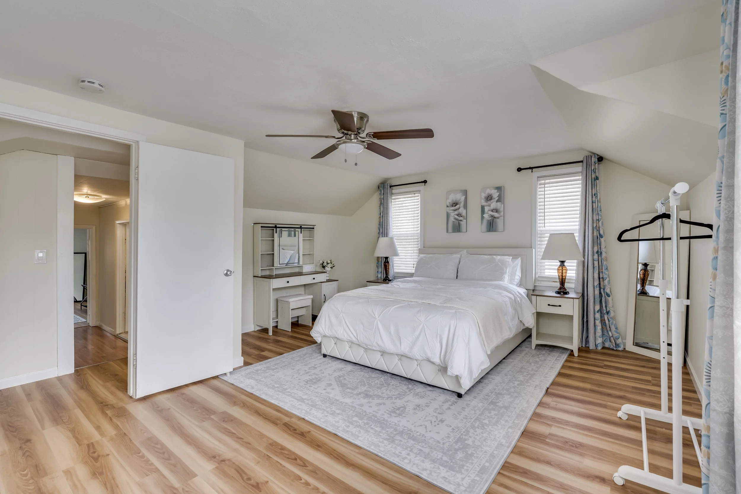 A bedroom with a white bed, nightstands, lamps, windows with blinds, a ceiling fan, a mirror, and a white clothing rack.