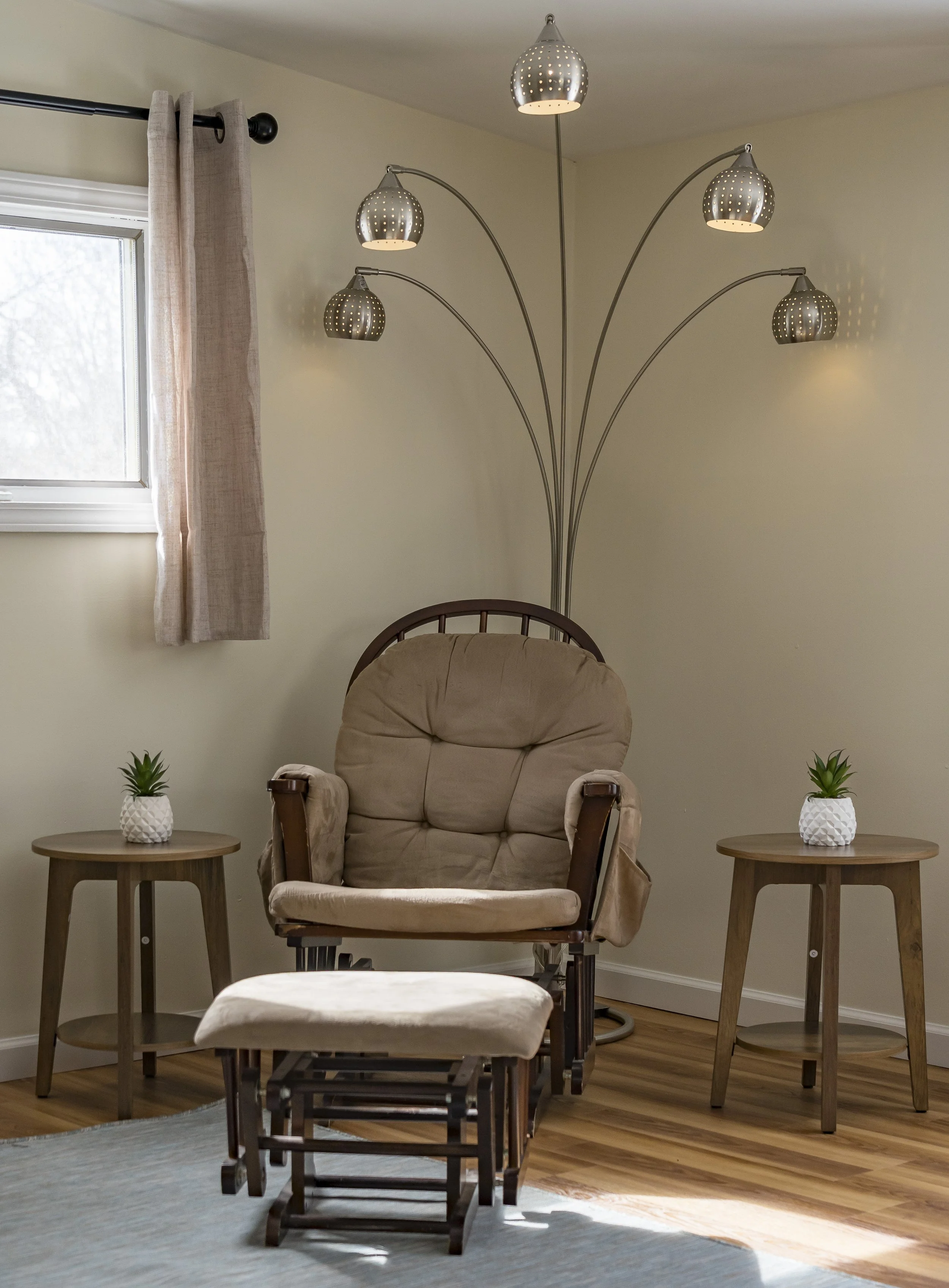 Comfortable beige armchair with a matching footstool, flanked by two wooden side tables each with a small potted plant, a modern floor lamp with five curved arms and perforated metallic shades, a window with beige curtains, on a light hardwood floor.