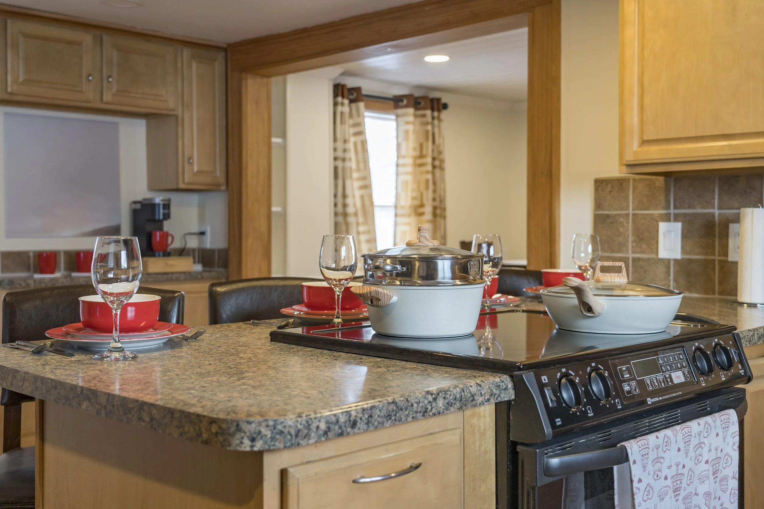 Kitchen with a granite countertop island, two white pots on an electric stove, three empty wine glasses, red bowls, and black chairs.