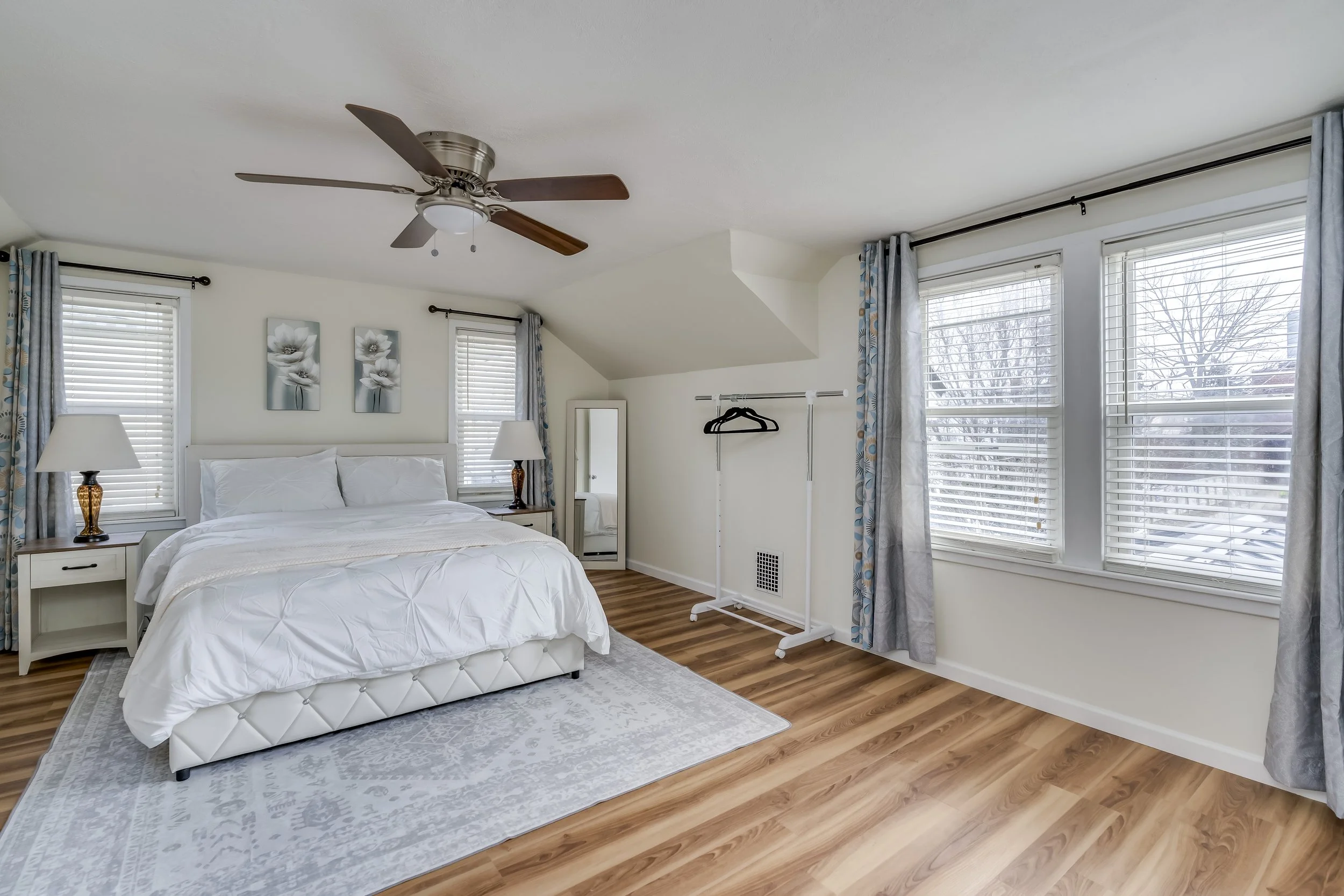 A bedroom with a white bed, two bedside tables with lamps, a standing mirror, and three large windows with curtains, hardwood floors, and a ceiling fan.