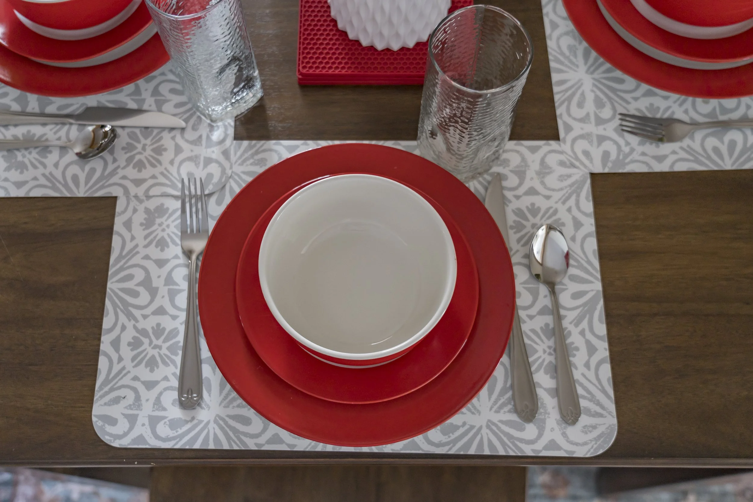 A table setting with a white and red plate, a white bowl, silver utensils, two tall water glasses, gray and white patterned placemats, and a wooden table with red and white dishes.