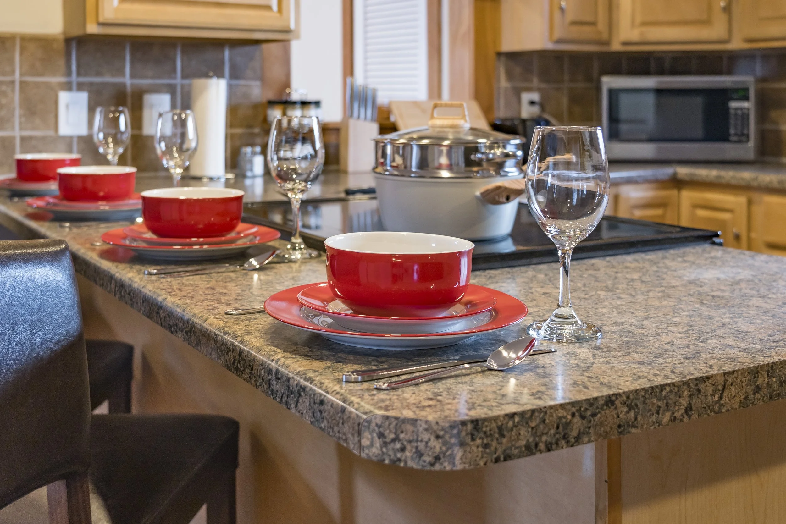 Kitchen island set for four with red bowls, matching red plates, clear wine glasses, and silverware, with a ceramic pot and a microwave in the background.