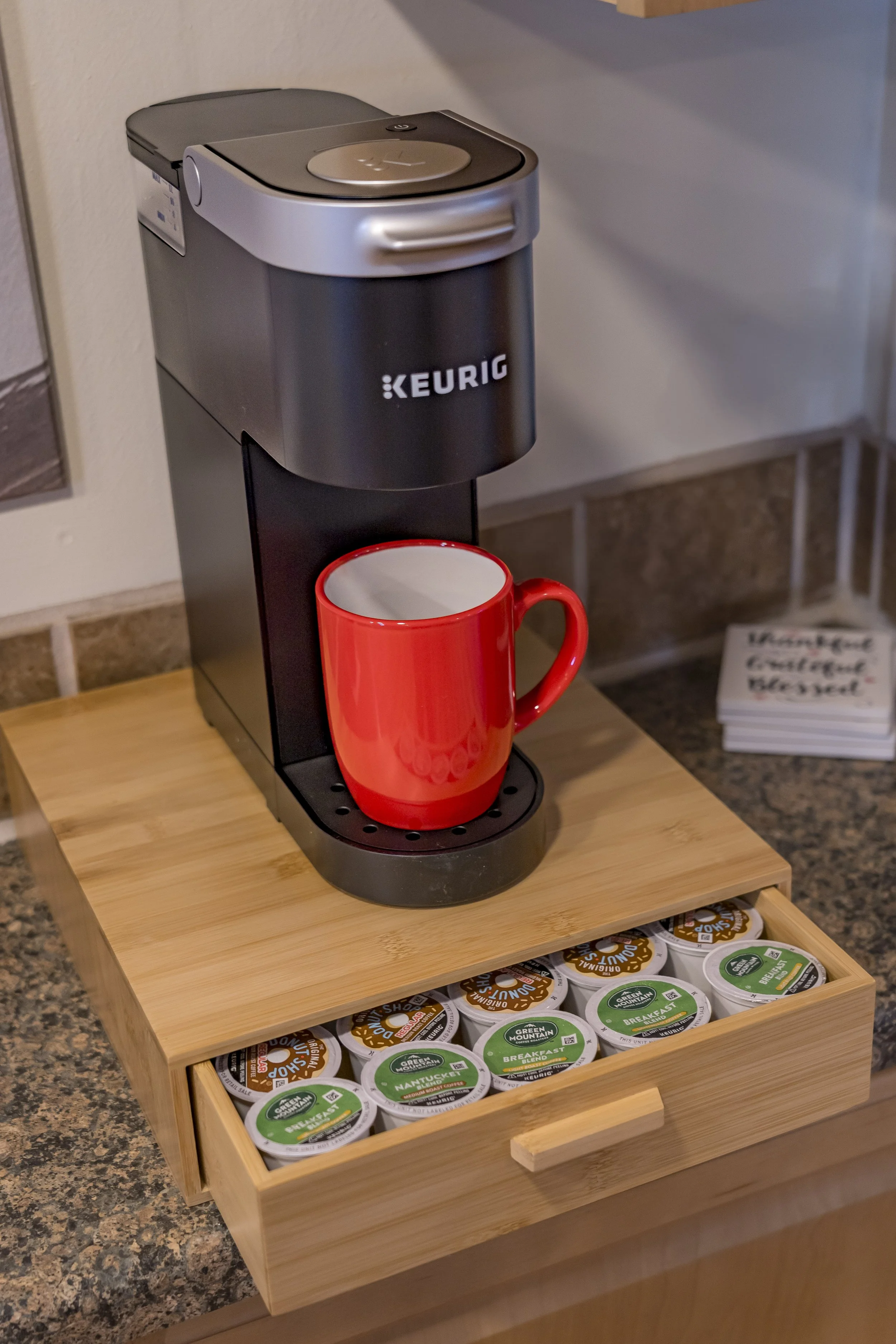 A Keurig coffee maker with a red mug on the drip tray, placed on a wooden tray with organized coffee pods, on a kitchen counter.
