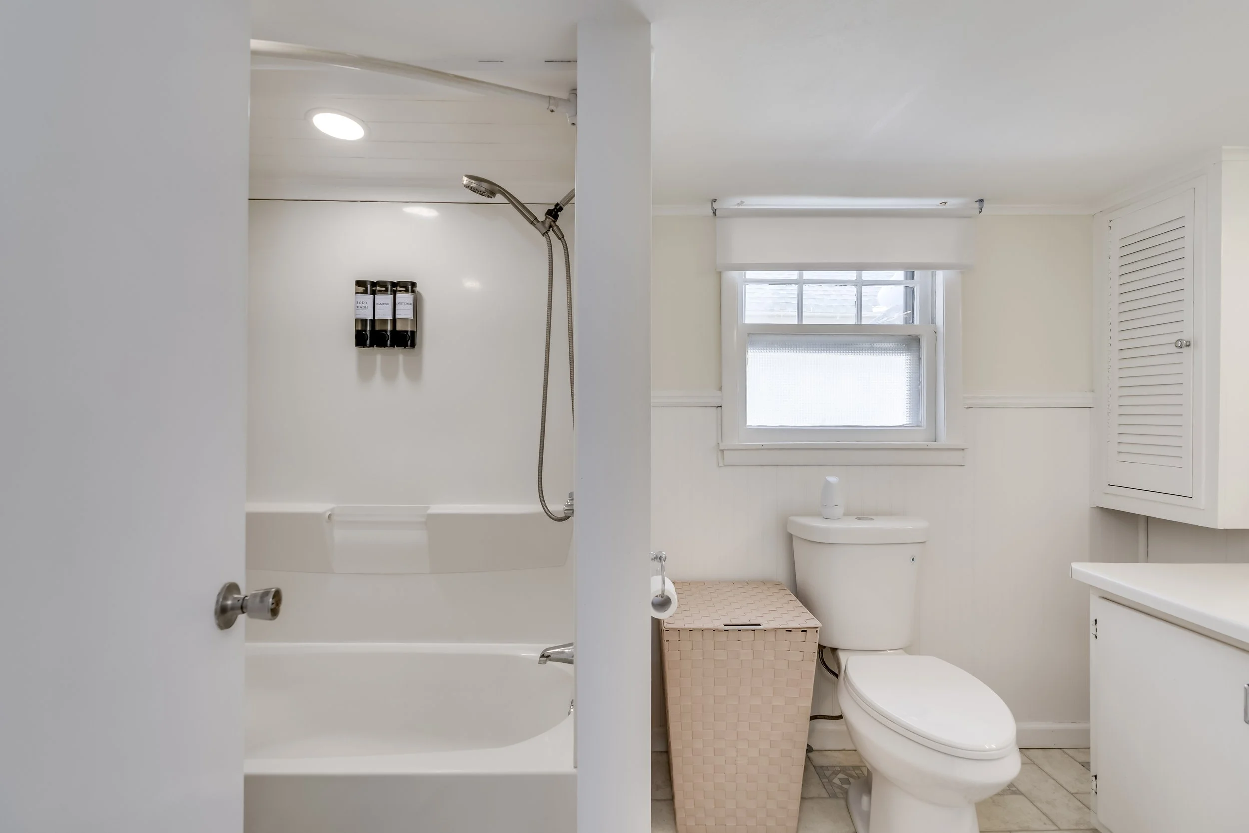 A small bathroom with a shower and a toilet, white walls, a window, and a beige laundry basket.