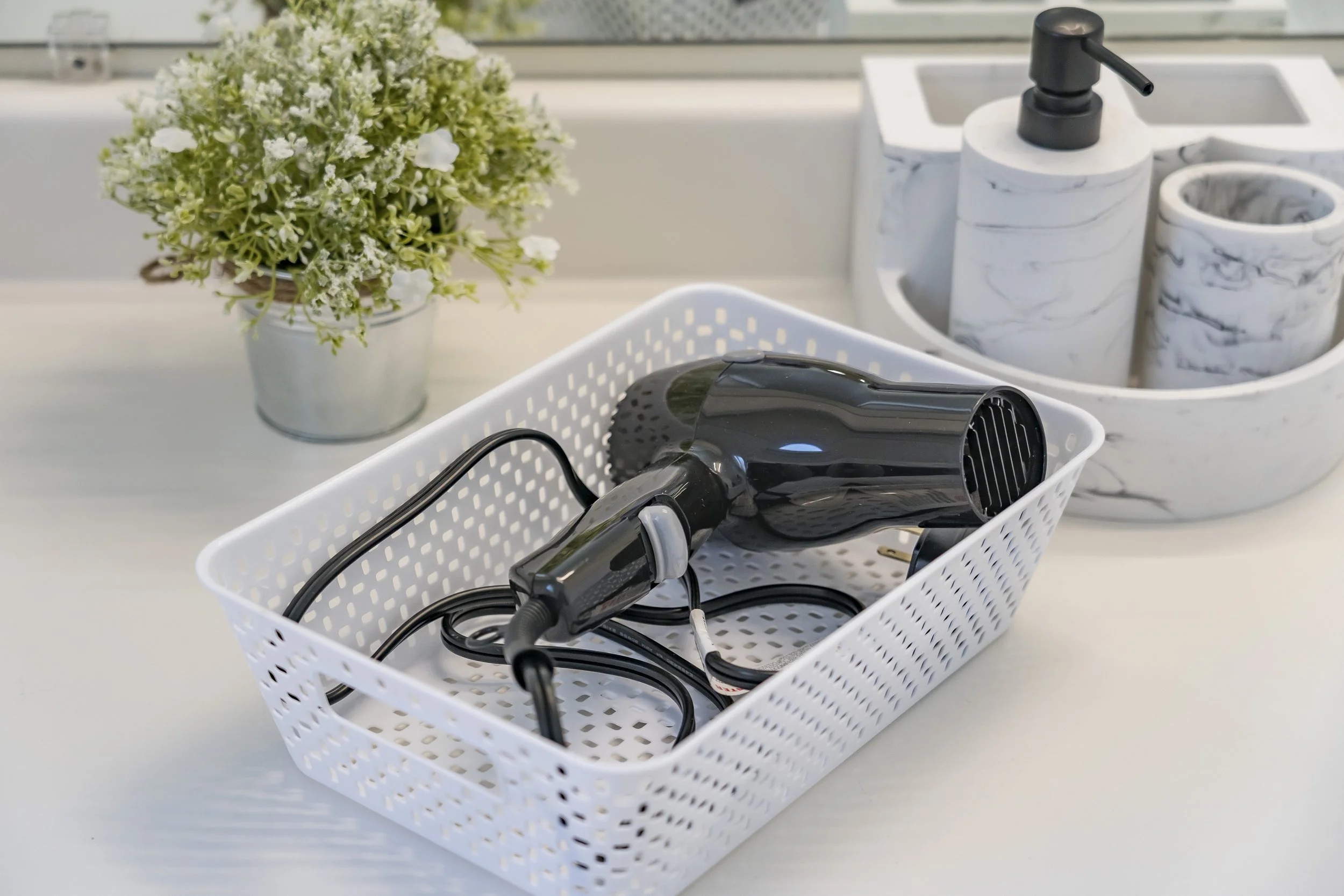Hair dryer and hair styling tools in a white basket on a countertop, with a potted flower and marble-patterned containers in the background.