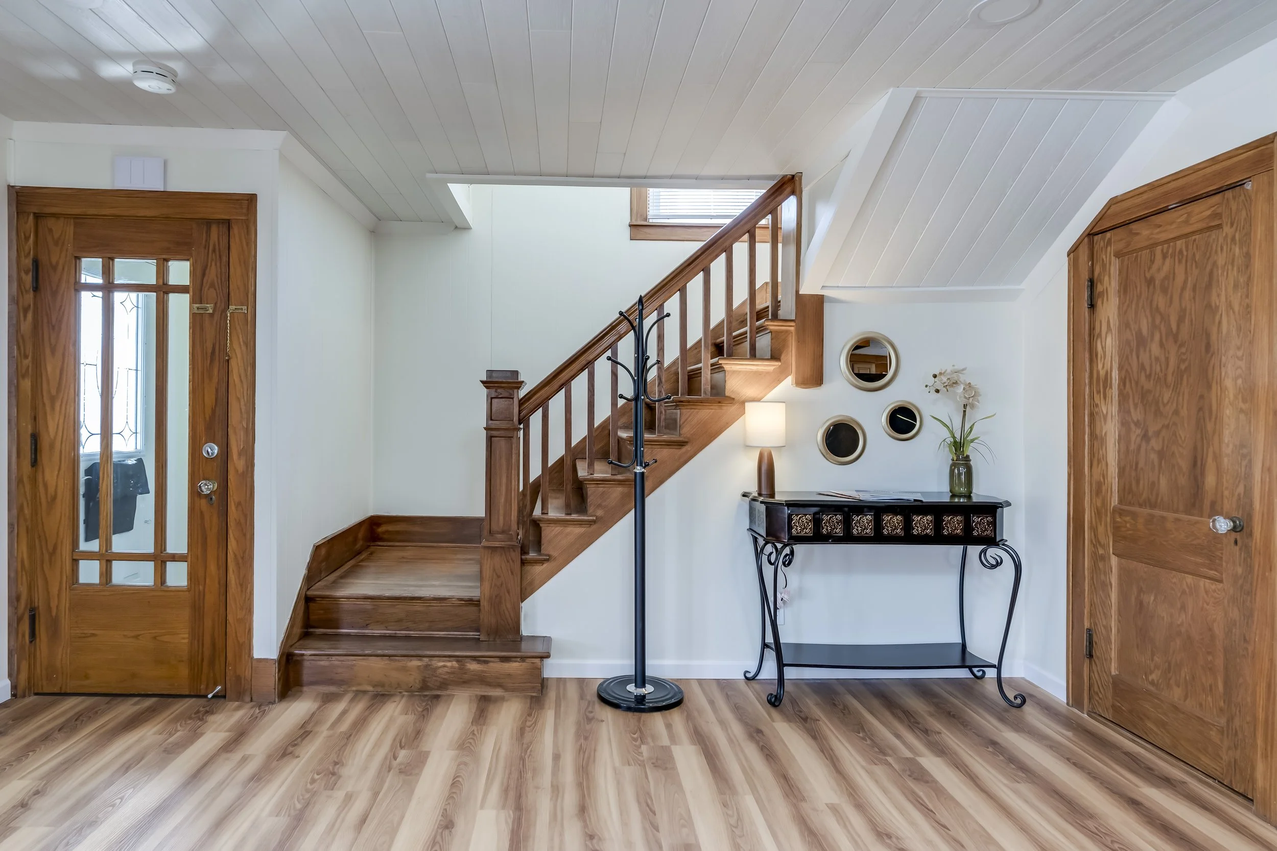 Interior of a house with wooden details and a staircase, showing a front door with glass panels, a small side table with decorative items, and wall mirrors.