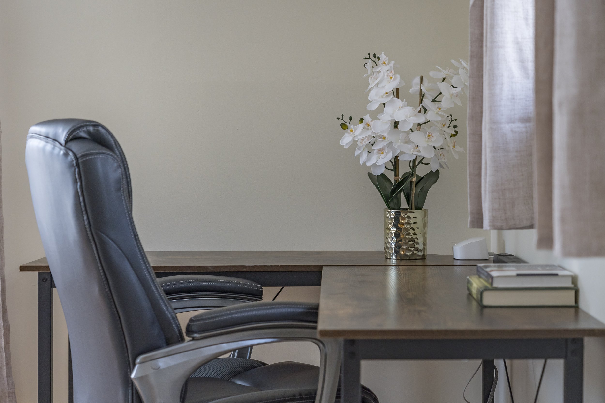 A wooden desk with a black leather office chair, a vase with white orchids, some books, a white speaker, and a lamp, with beige curtains hanging beside the desk.
