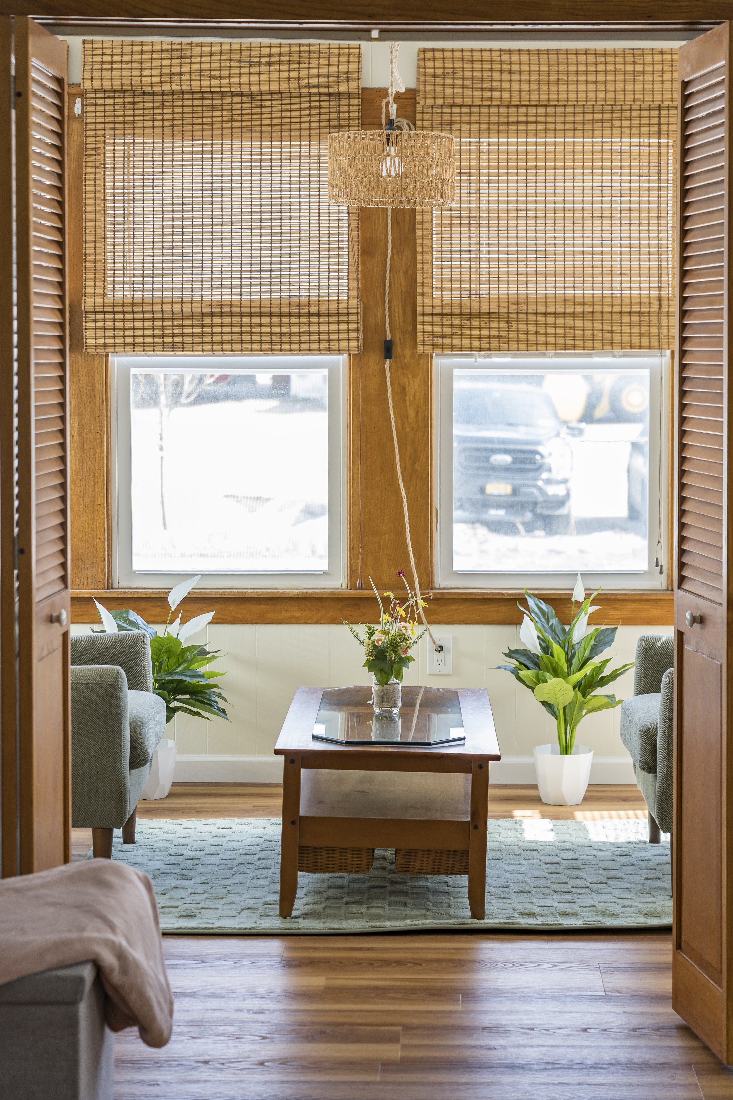 A cozy living room view through an open door, featuring a small wooden table with a flower arrangement, two armchairs, large potted plants, wooden window shutters, bamboo blinds, a woven pendant light, and a light-colored area rug on a wood floor.
