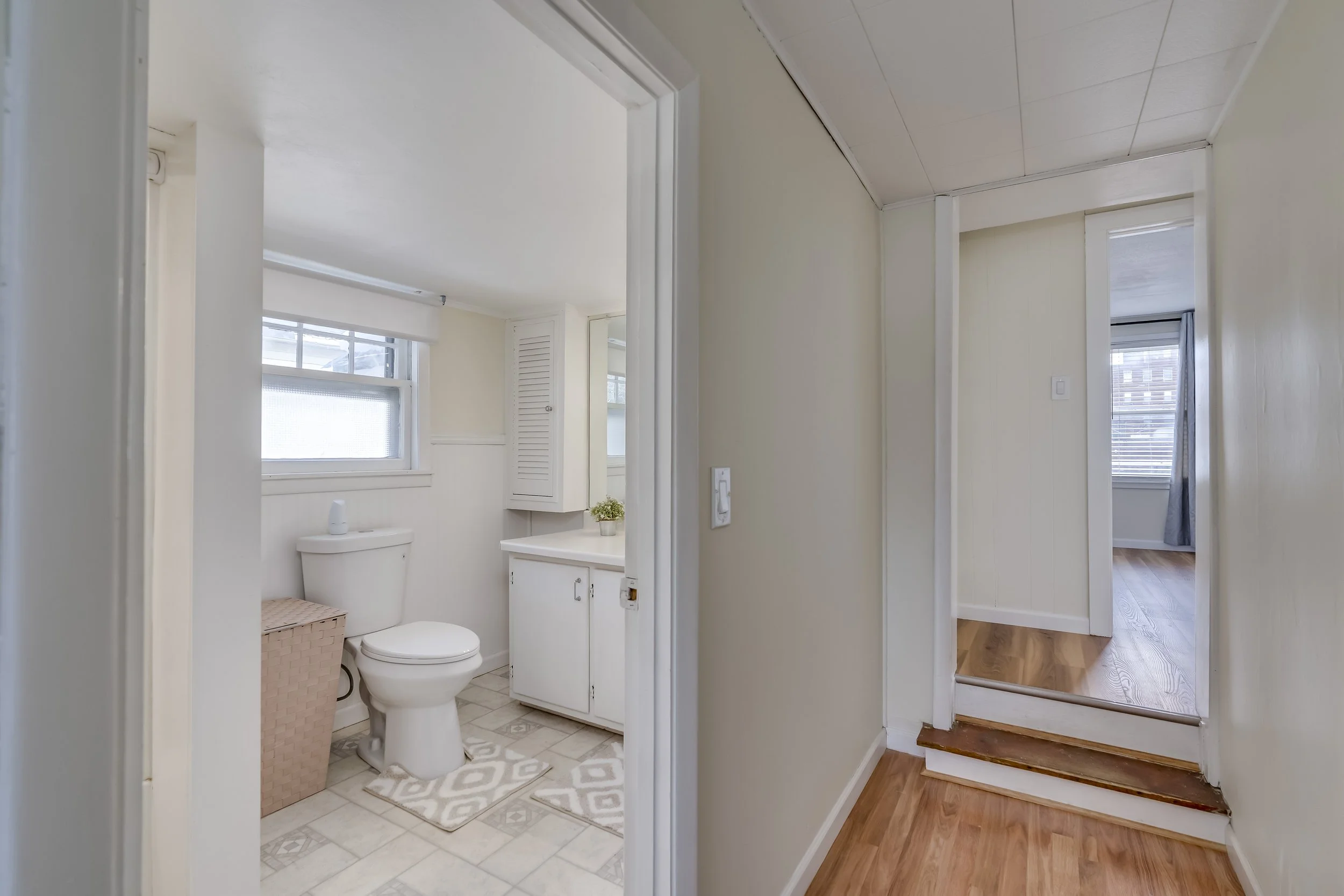 Interior view showing a small bathroom with a toilet, window, storage cabinet, and laundry basket on the tiled floor, and a hallway with wood flooring leading to a room with a window and curtain.