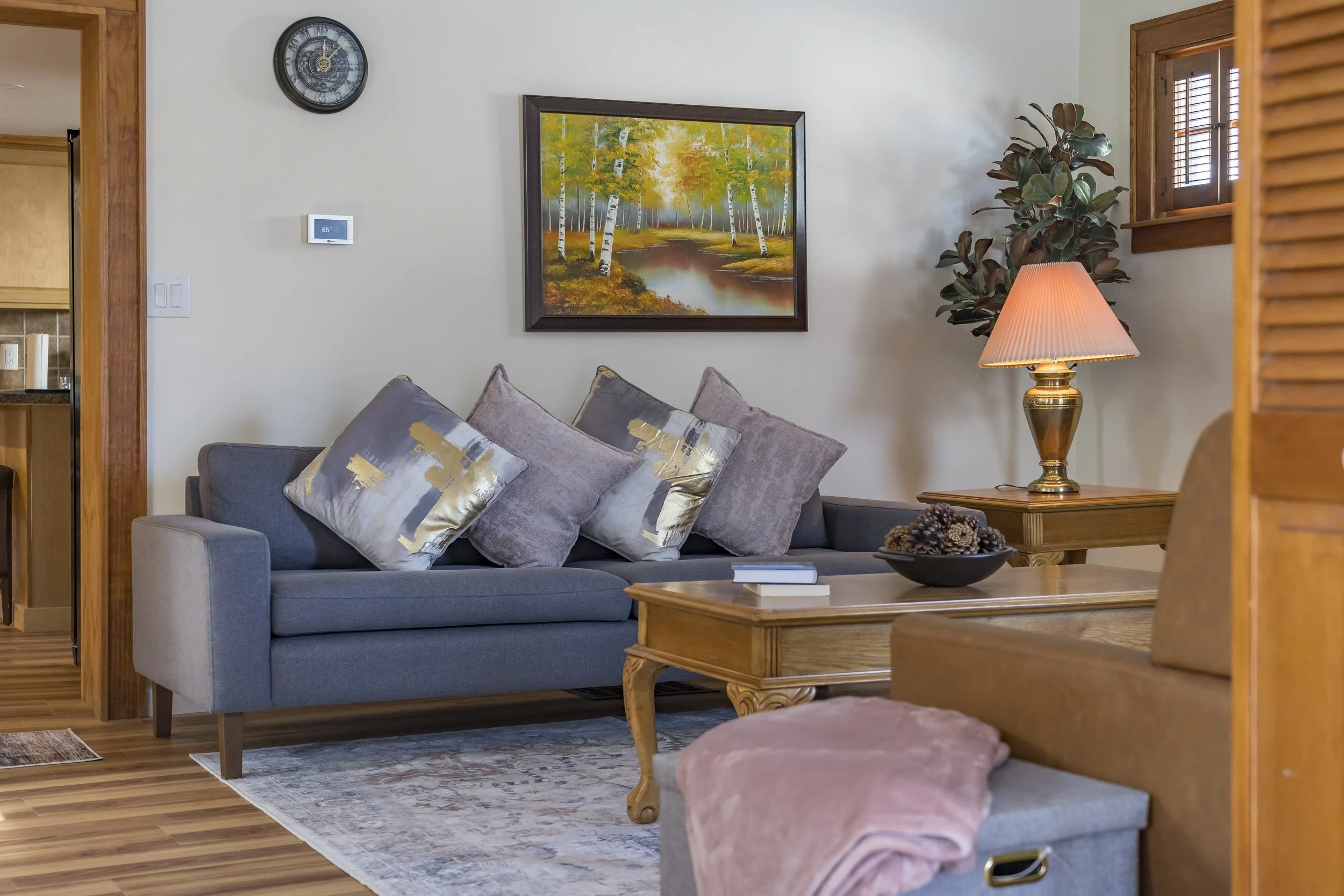Living room with a gray sofa decorated with purple and gold pillows, a wooden coffee table with a book and a decorative shell bowl, a table lamp with a pink shade, a framed painting of trees and a stream, a decorative plant, a wall clock, and a small