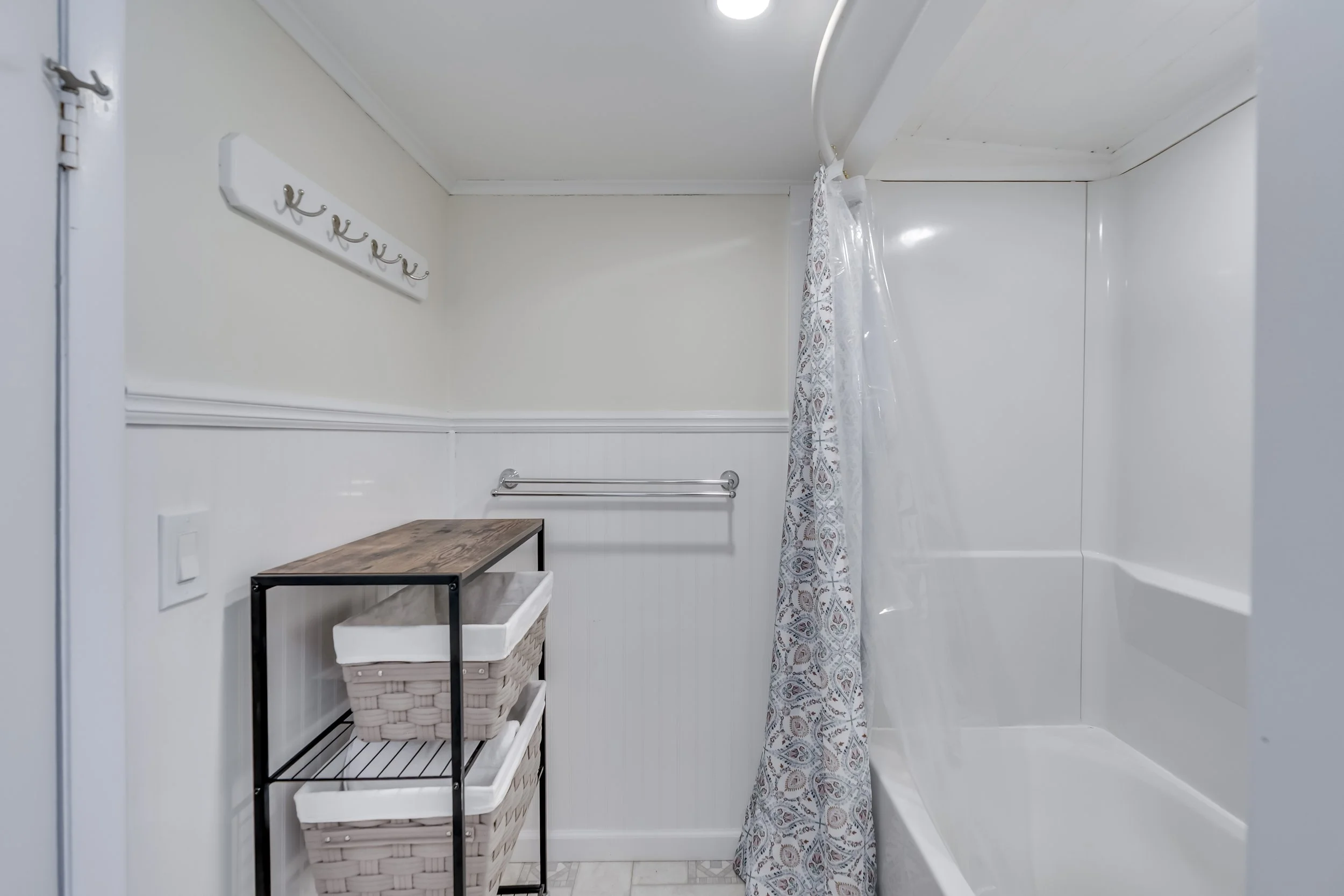 A small bathroom with a bathtub, shower curtain, wall hooks, a shelving unit with baskets, towel bar, and a decorative wall hook set.
