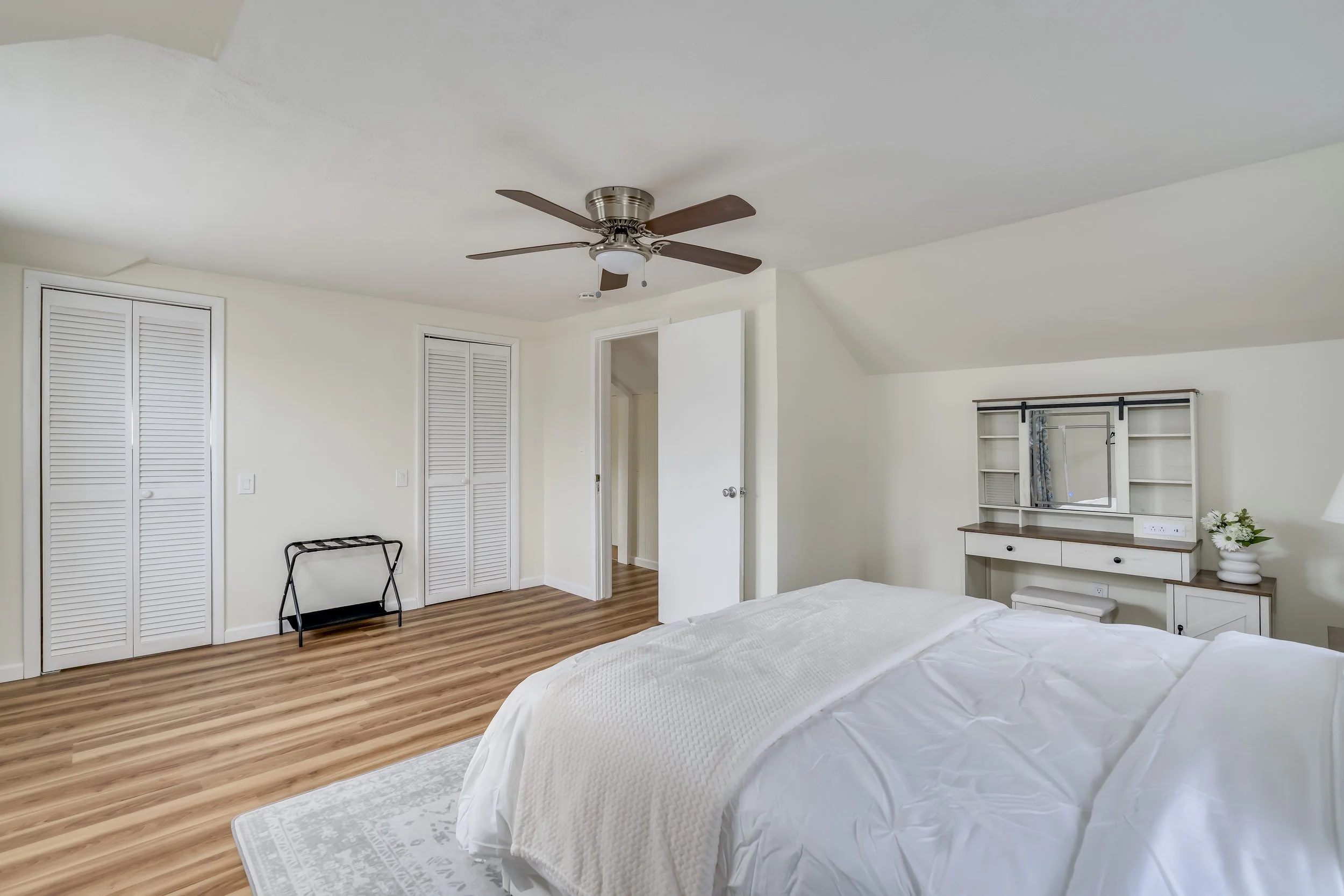 A bedroom with a white bed, wooden flooring, and a ceiling fan. Contains a dresser with a mirror, a small stool, a flower vase, and closed closet doors.