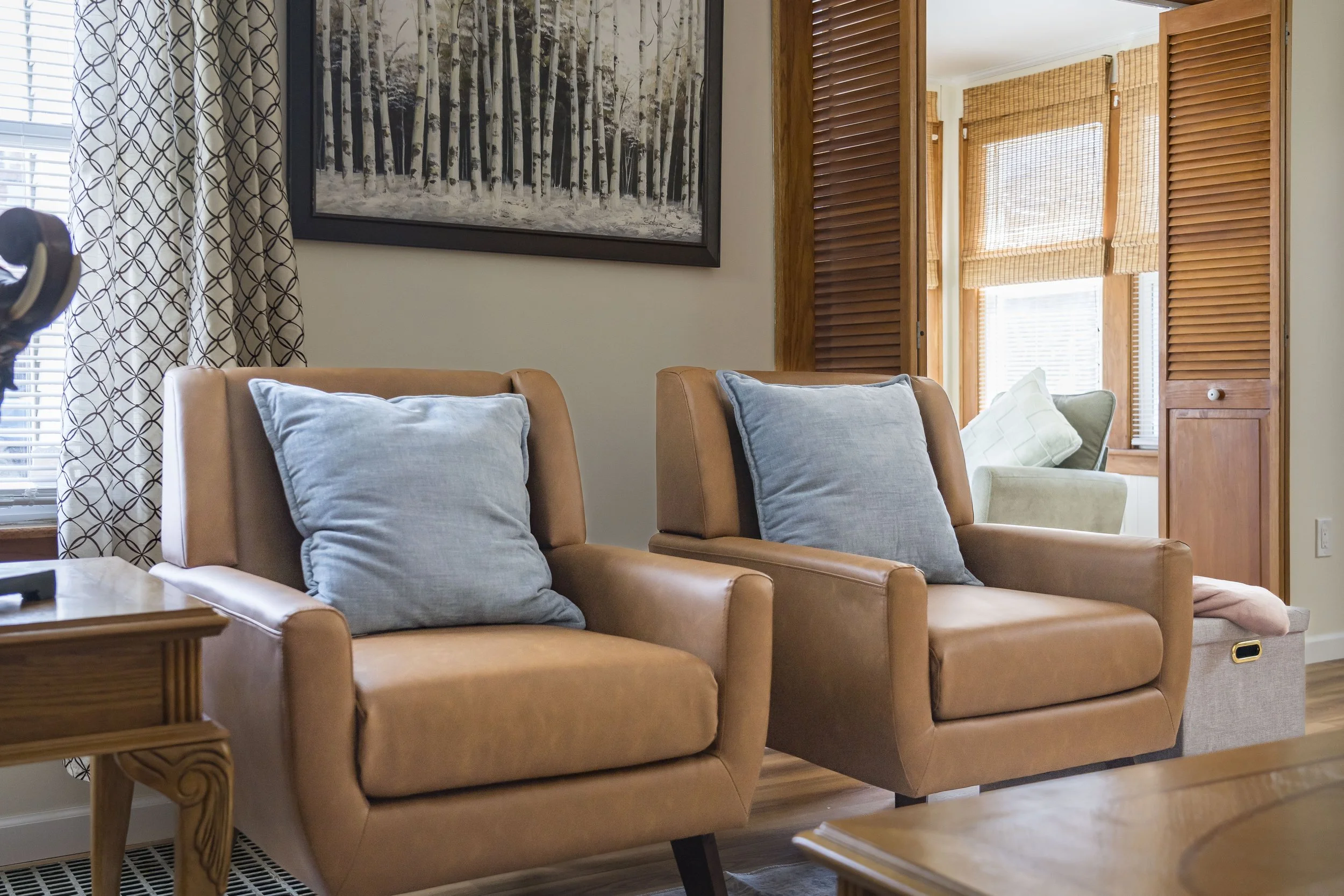 Living room with two tan armchairs, each with a light blue pillow, wooden side table, window with patterned curtains, and a painted forest scene on the wall. In the background, there is a sunroom with wooden blinds and a cushioned chair.