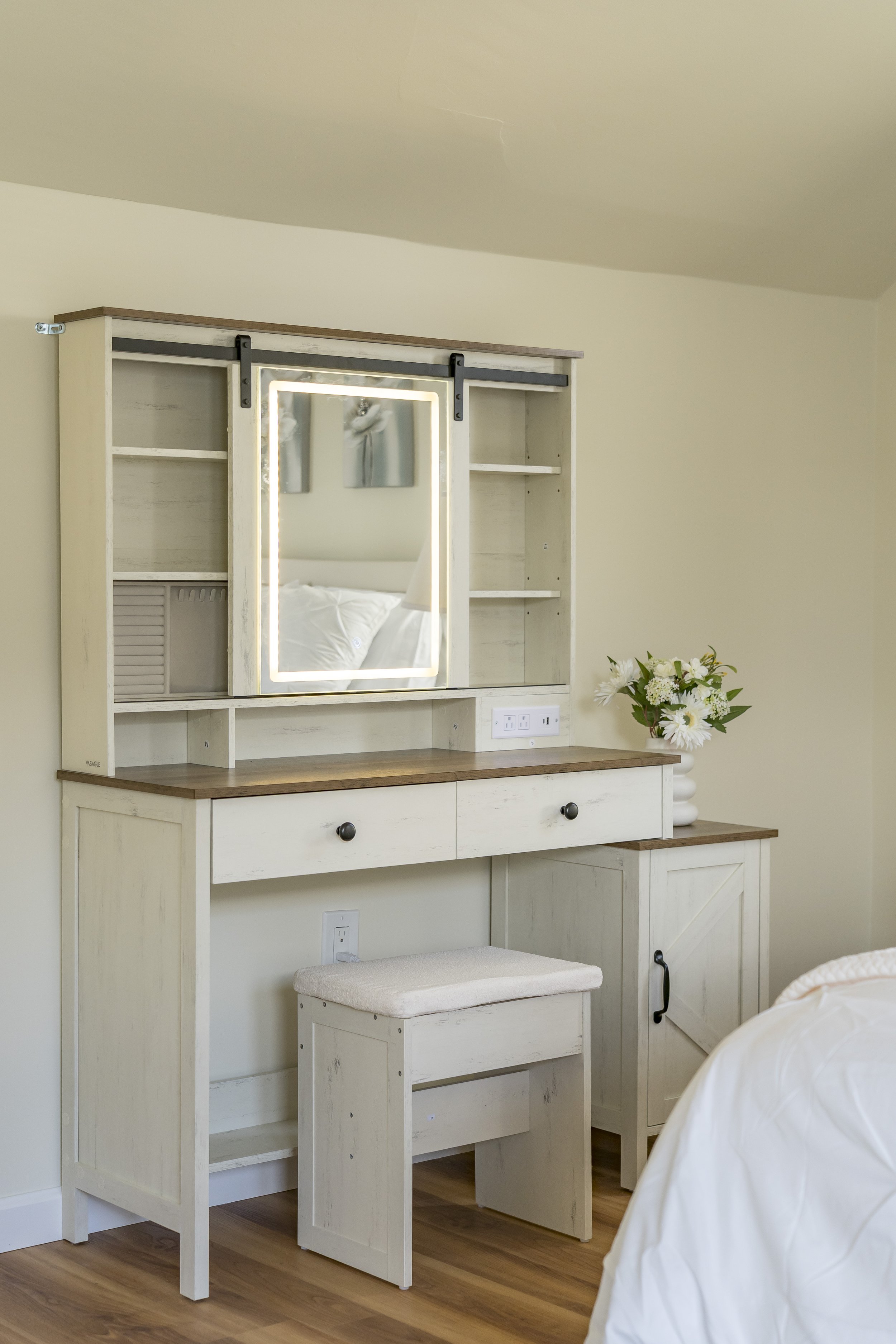 A bedroom vanity with a mirror and drawers, decorated with white flowers in a vase, set against a wall with electrical outlets.