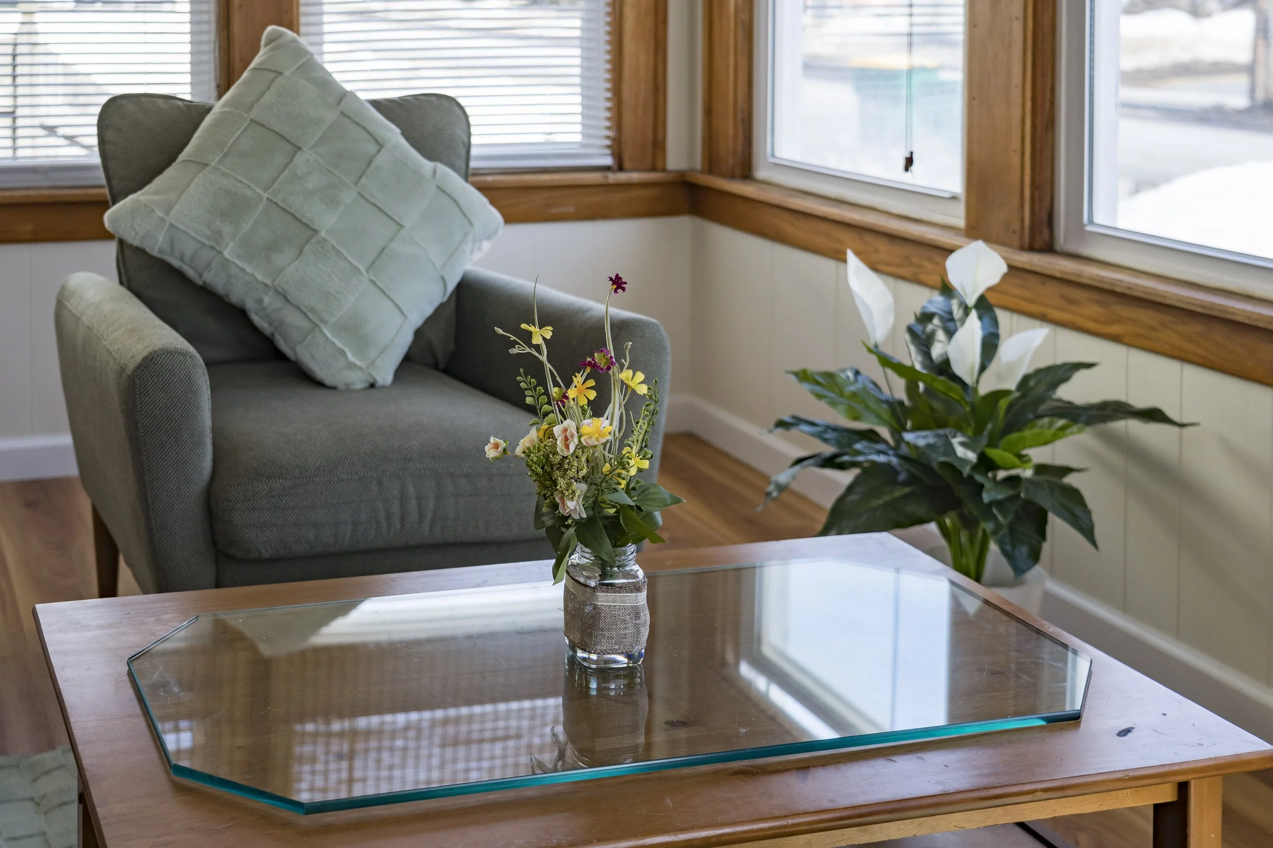 Living room corner with a gray armchair, a large pale green pillow, a wooden coffee table with a flower arrangement in a glass jar, and a potted peace lily plant, near windows with wooden frames and blinds.
