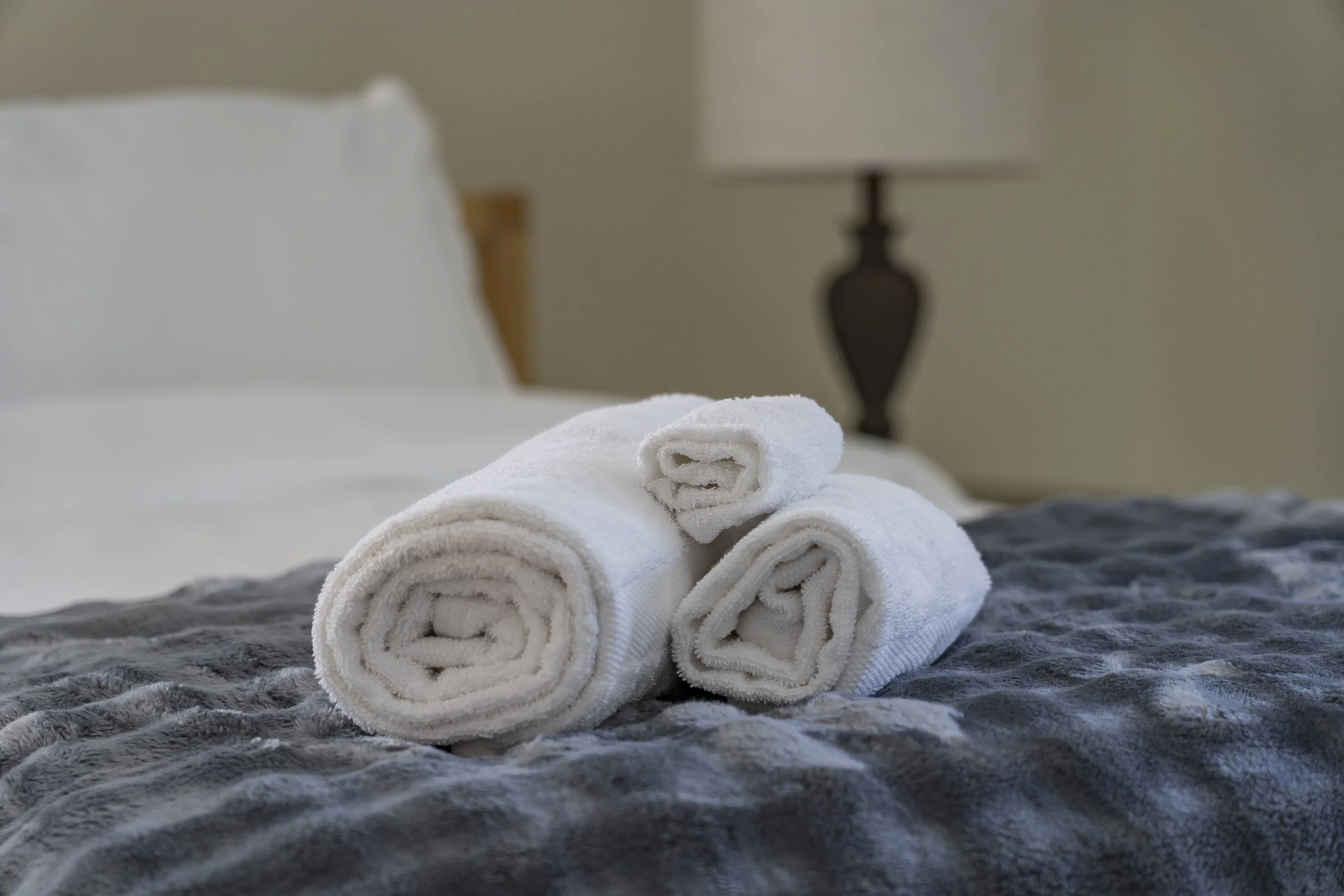 Three rolled white towels on a bed with a plush gray blanket, a white pillow, a wooden headboard, and a dark brown bedside lamp in a neutral-colored bedroom.