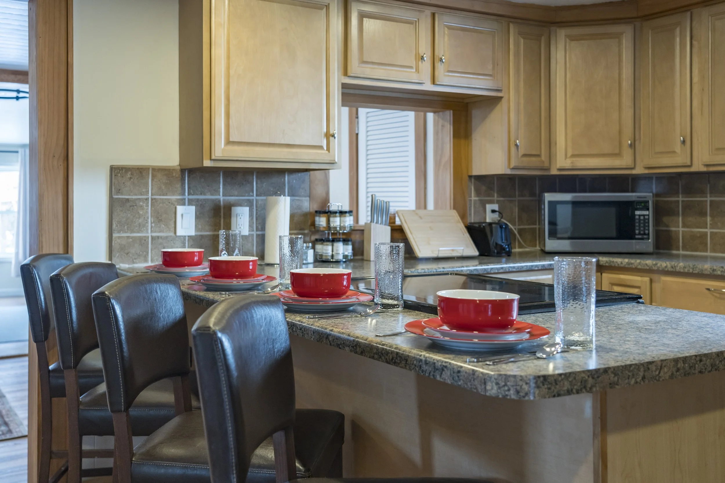 Kitchen with a granite island counter set for four, featuring red bowls and plates, tall drinking glasses, and silverware. Black chairs surround the island. The background includes wooden cabinets, a microwave, and various kitchen items.