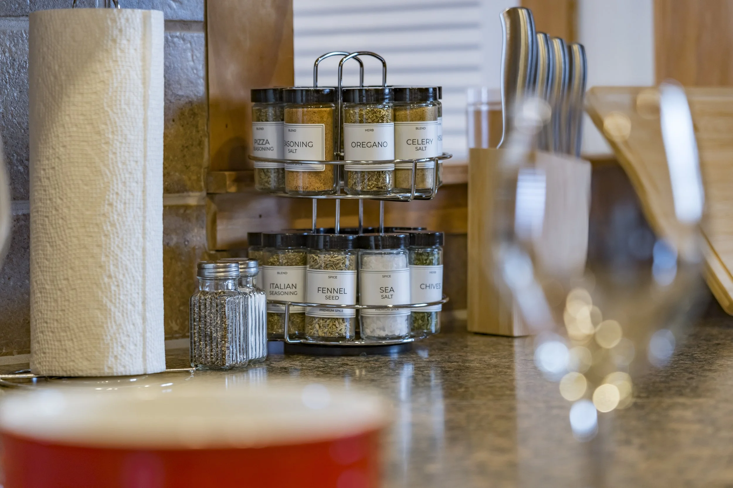 Kitchen countertop with spice jars on a two-tiered metal rack, small glass jar with pepper, paper towel roll, and utensils in a holder.