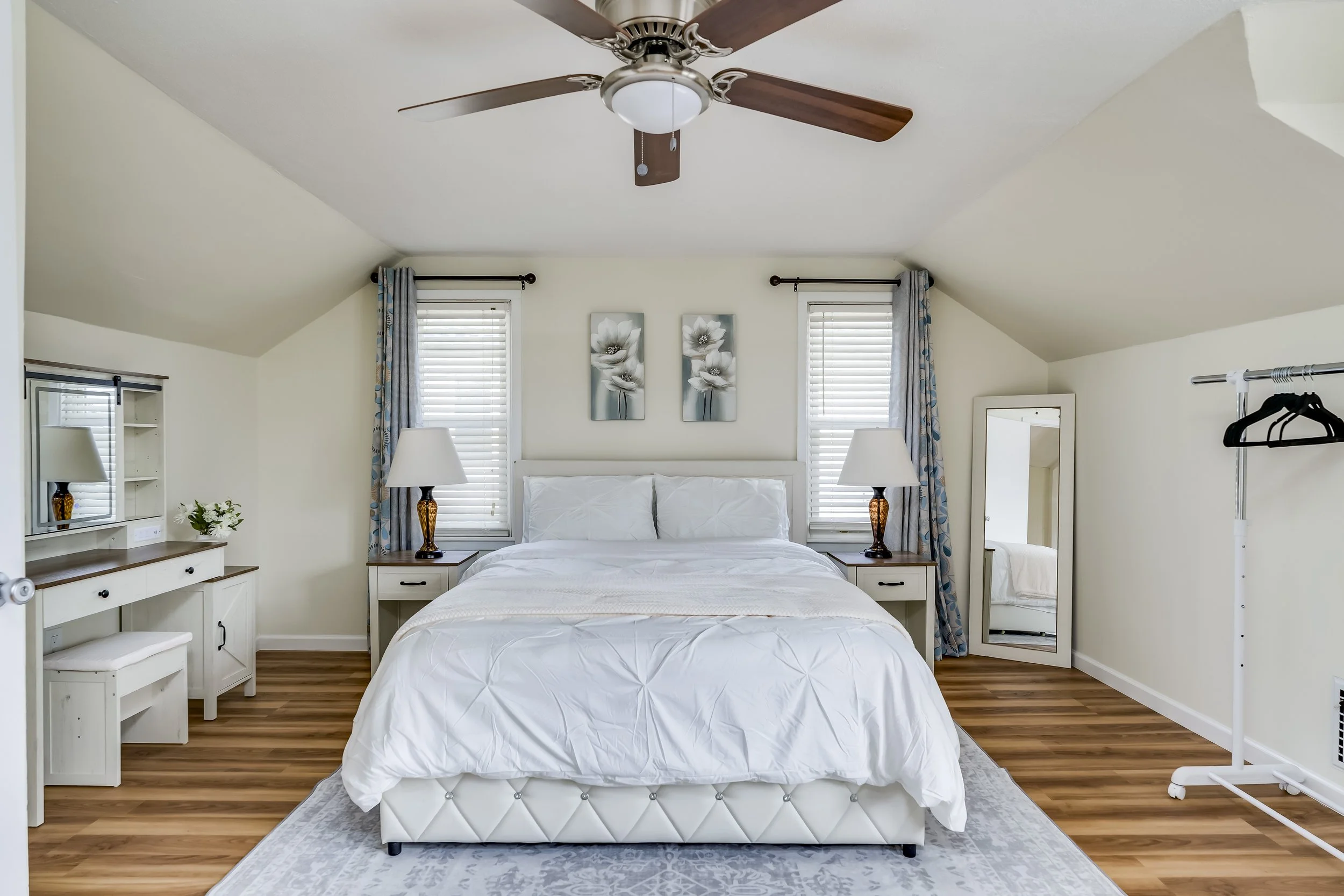 A bedroom with a white bed, two windows with blue and patterned curtains, two bedside tables with lamps, a large mirror, a vanity with a chair, and a clothing rack, all with a ceiling fan overhead.