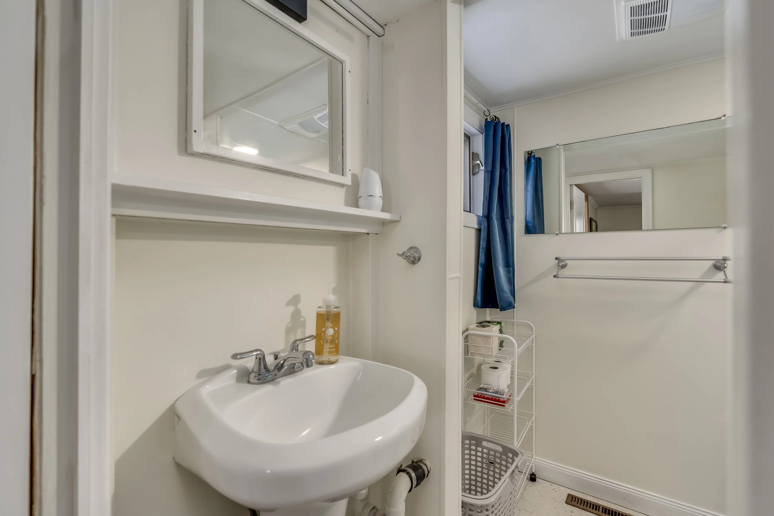 Small bathroom with white walls, a utility sink with a soap dispenser on it, a small mirror above the sink, a window with blue curtains, a large mirror, a towel bar, a metal storage rack with toilet paper and books, and a floor vent.