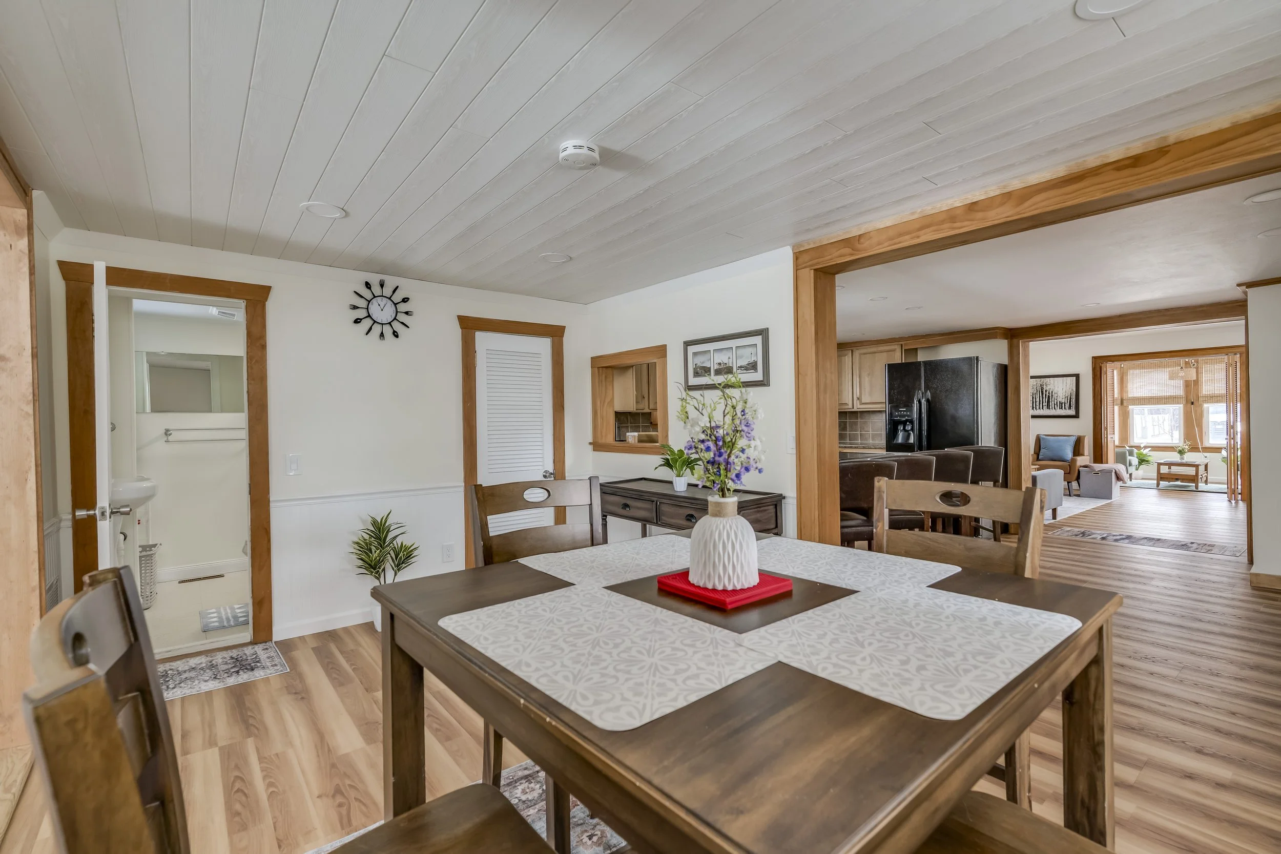 Dining room with a wooden table, flowers in a white vase, and view into a living room area with a black refrigerator, sofa, and windows.